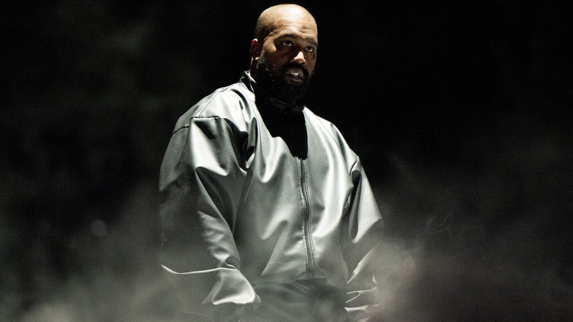 Ye in a dark, oversized jacket, standing amidst a foggy, dimly lit background.