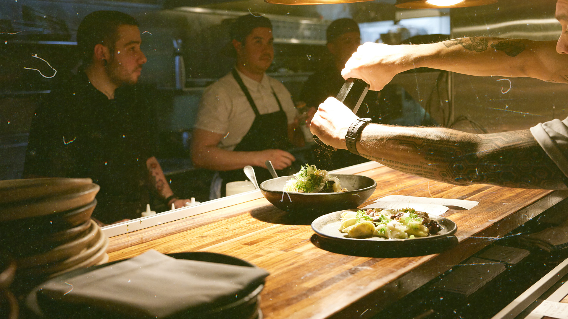 Chefs in a dimly lit kitchen prepare and garnish dishes on a wooden counter, with stacks of plates nearby.