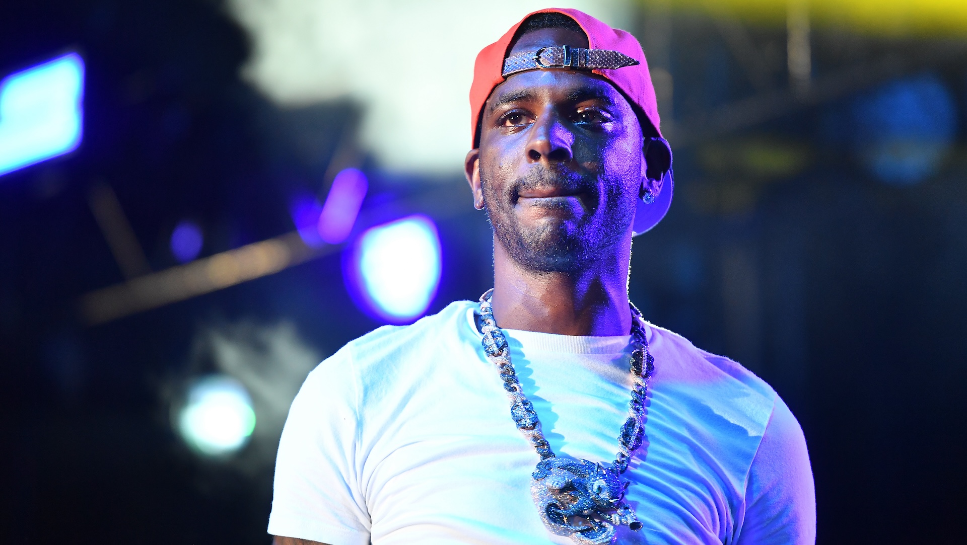 Young Dolph wearing a white t-shirt, red cap, and a large chain necklace on stage with colorful lights in the background.