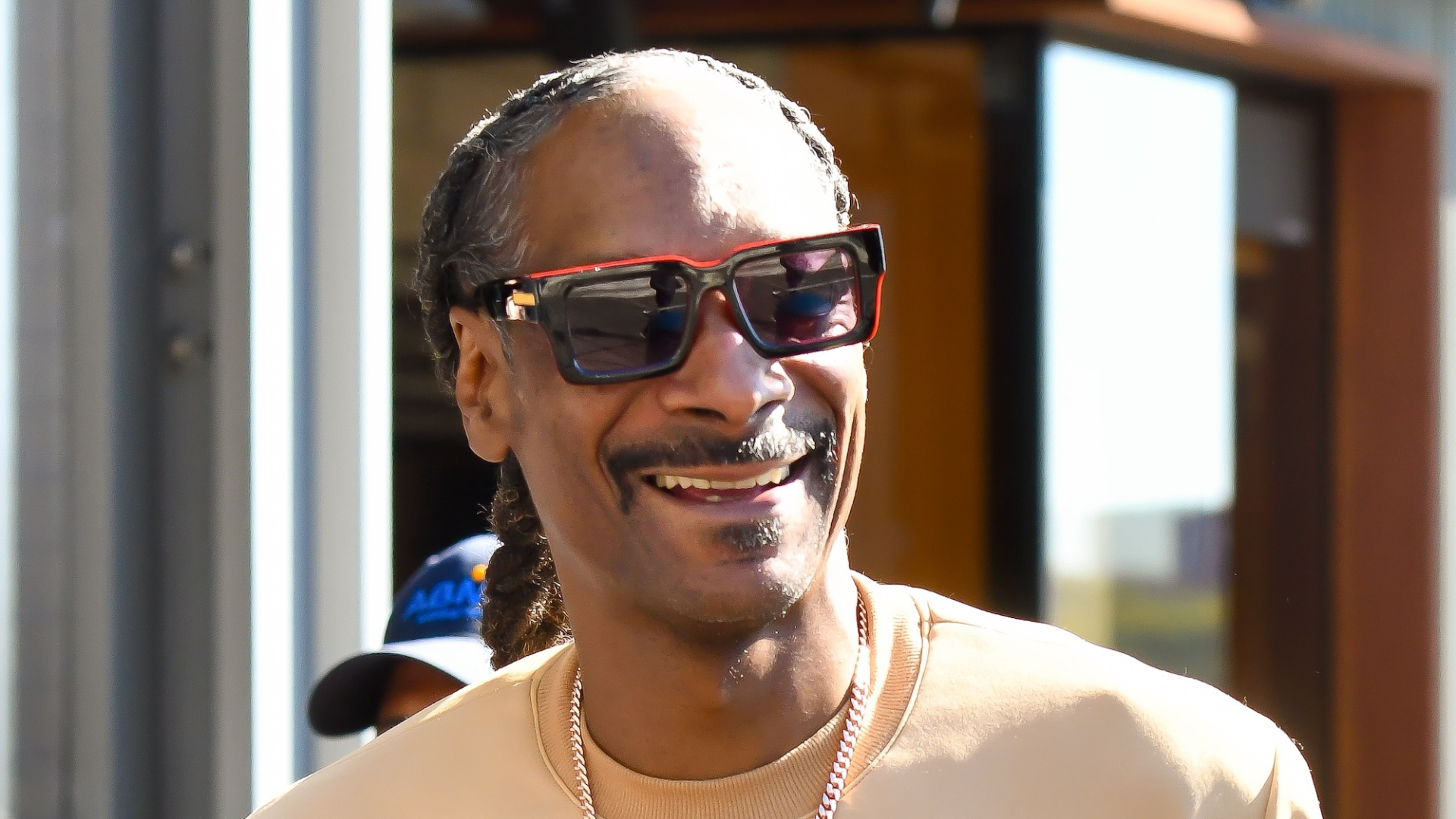 Snoop Dogg smiling, wearing sunglasses and a beige shirt, with braided hair.