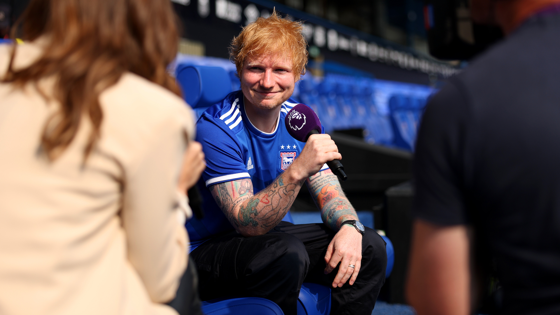 Ed Sheeran, wearing an Ipswich Town jersey, sits on a stadium bench holding a microphone, engaged in an interview.