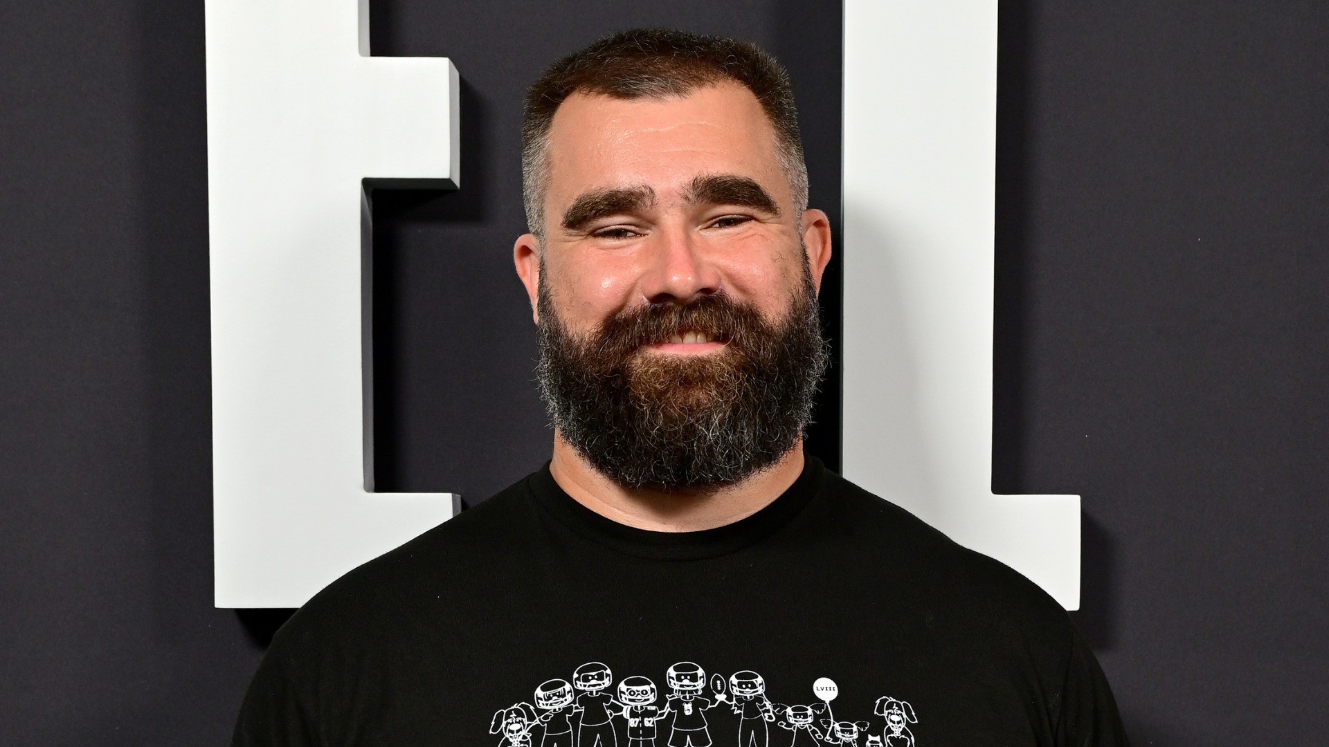 A man with a thick beard and short hair smiles, wearing a black t-shirt with a cartoon design, standing against a dark background.