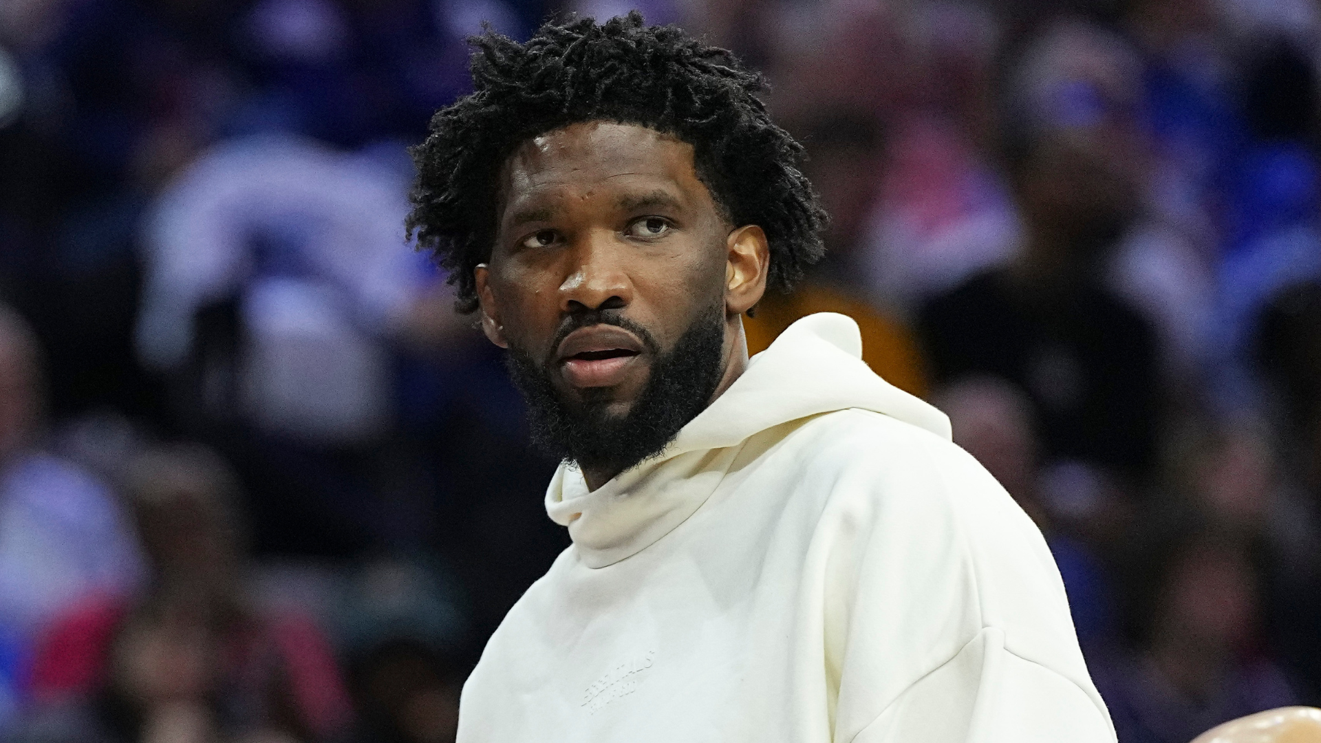 A man with a beard and curly hair wearing a white hoodie, standing in a crowd.