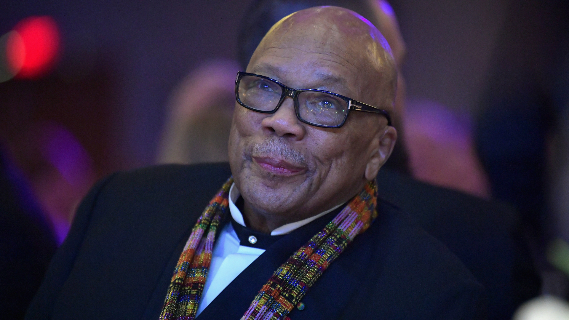 Quincy Jones, wearing glasses and a colorful scarf, smiles at an event.
