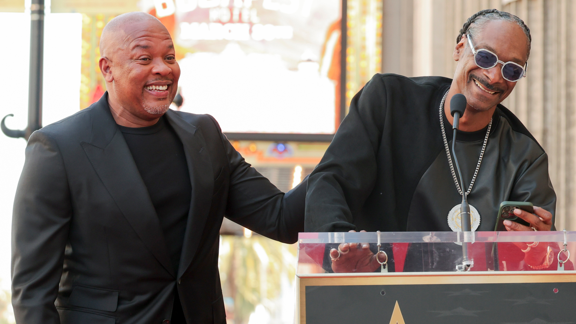 Dr. Dre and Snoop Dogg smiling at a podium event. Dr. Dre is wearing a black suit, and Snoop Dogg is in sunglasses and a black outfit.