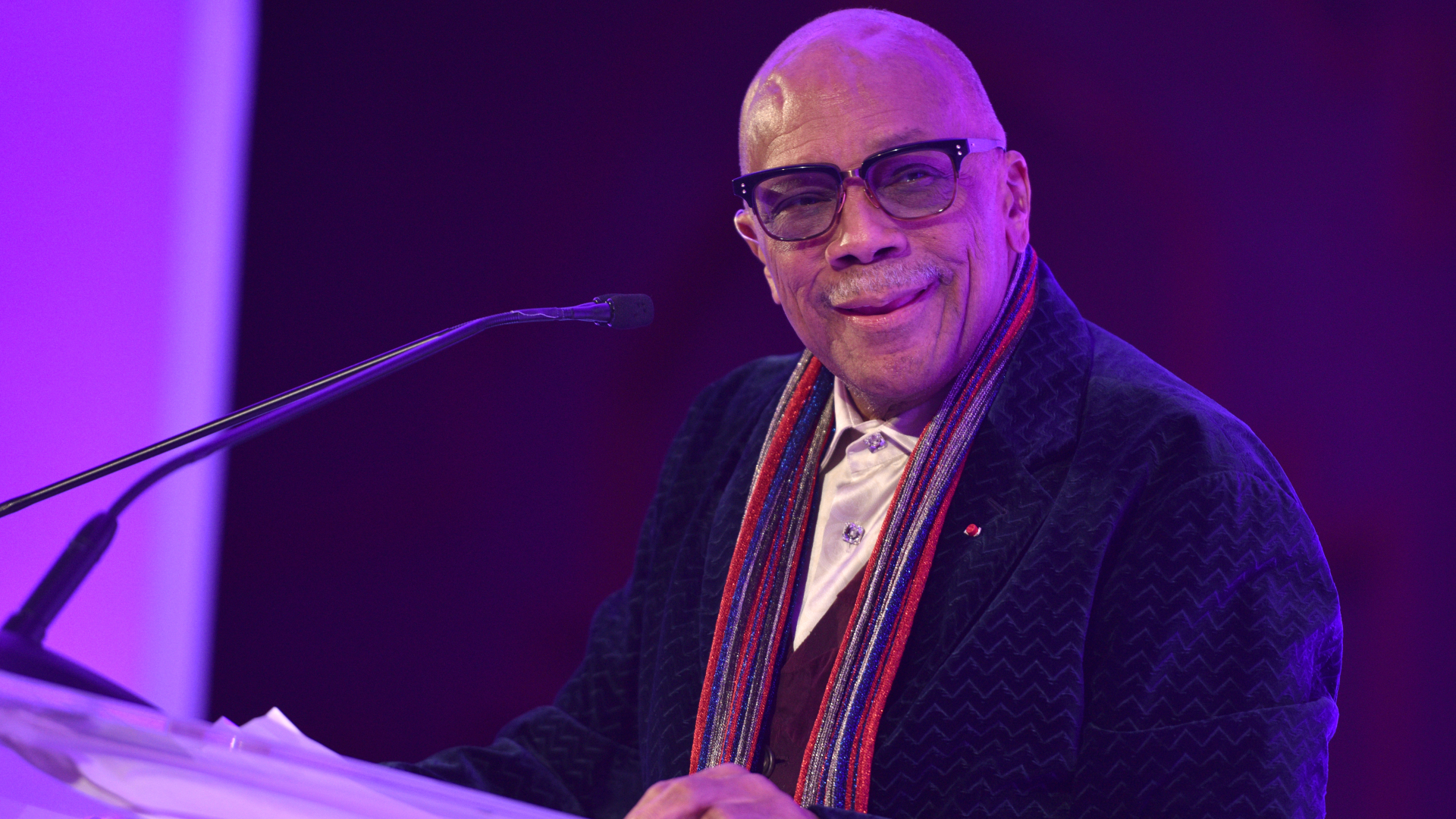 Quincy Jones, wearing glasses and a colorful scarf, stands at a podium speaking.