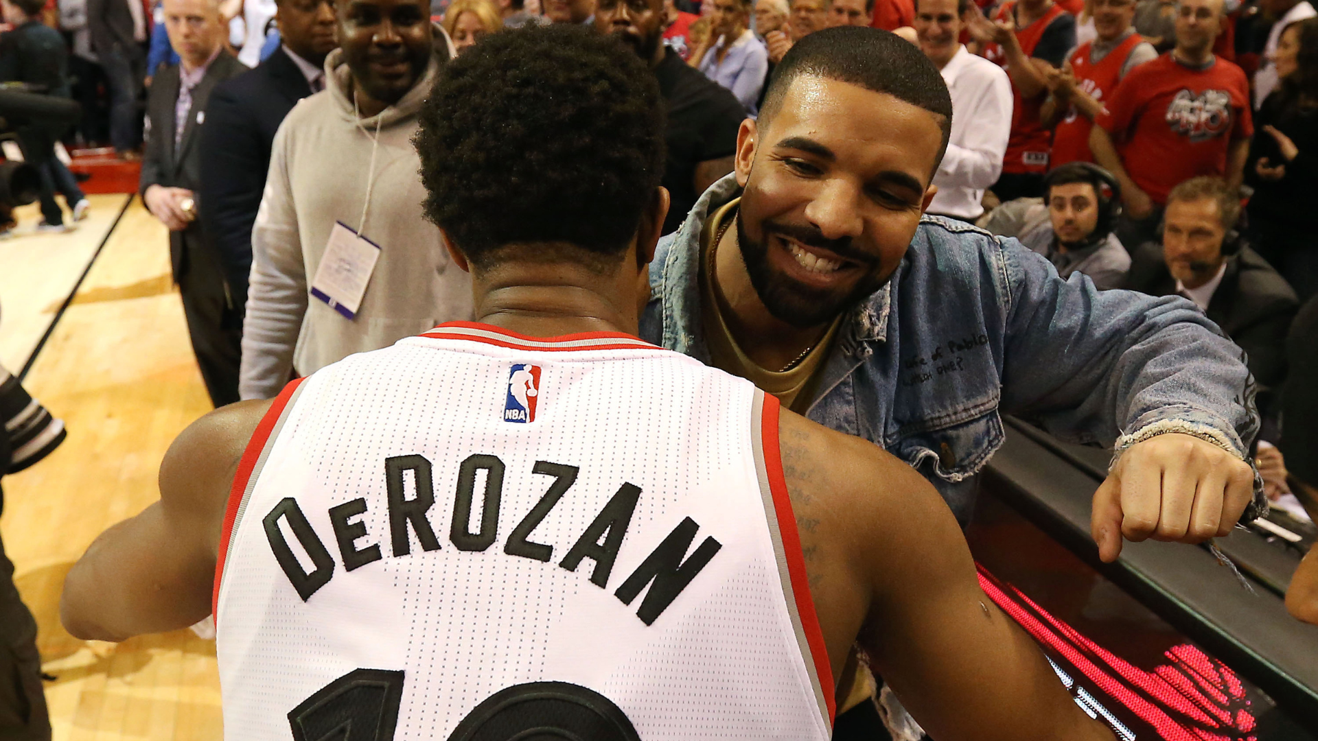 Drake smiling and greeting DeMar DeRozan, wearing a Toronto Raptors jersey, at a basketball game.