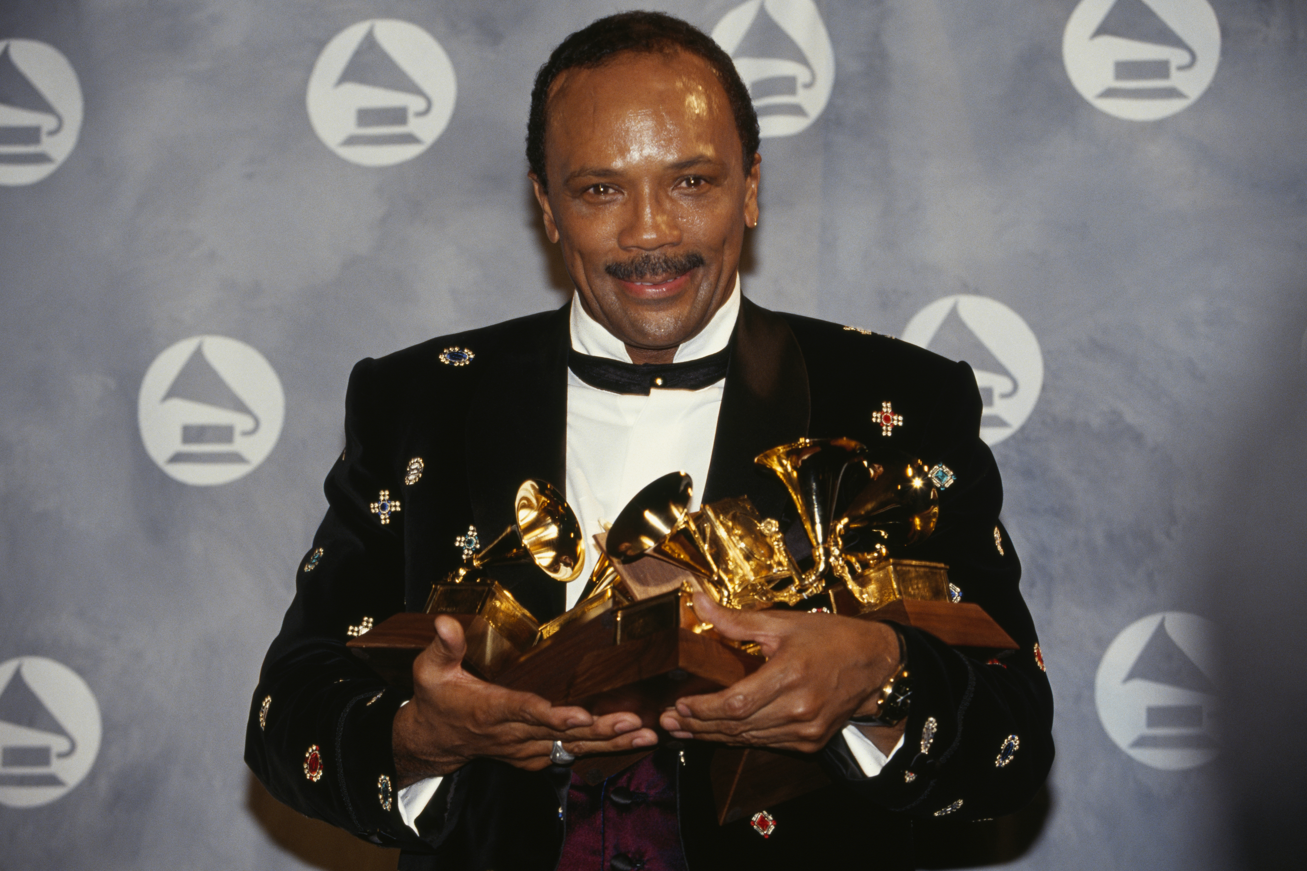 Quincy Jones holding multiple Grammy Awards, smiling, in a formal outfit with a patterned jacket.