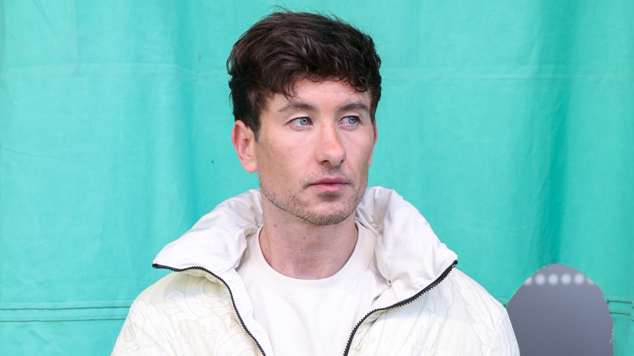 Barry Keoghan wearing a white jacket stands against a turquoise background.