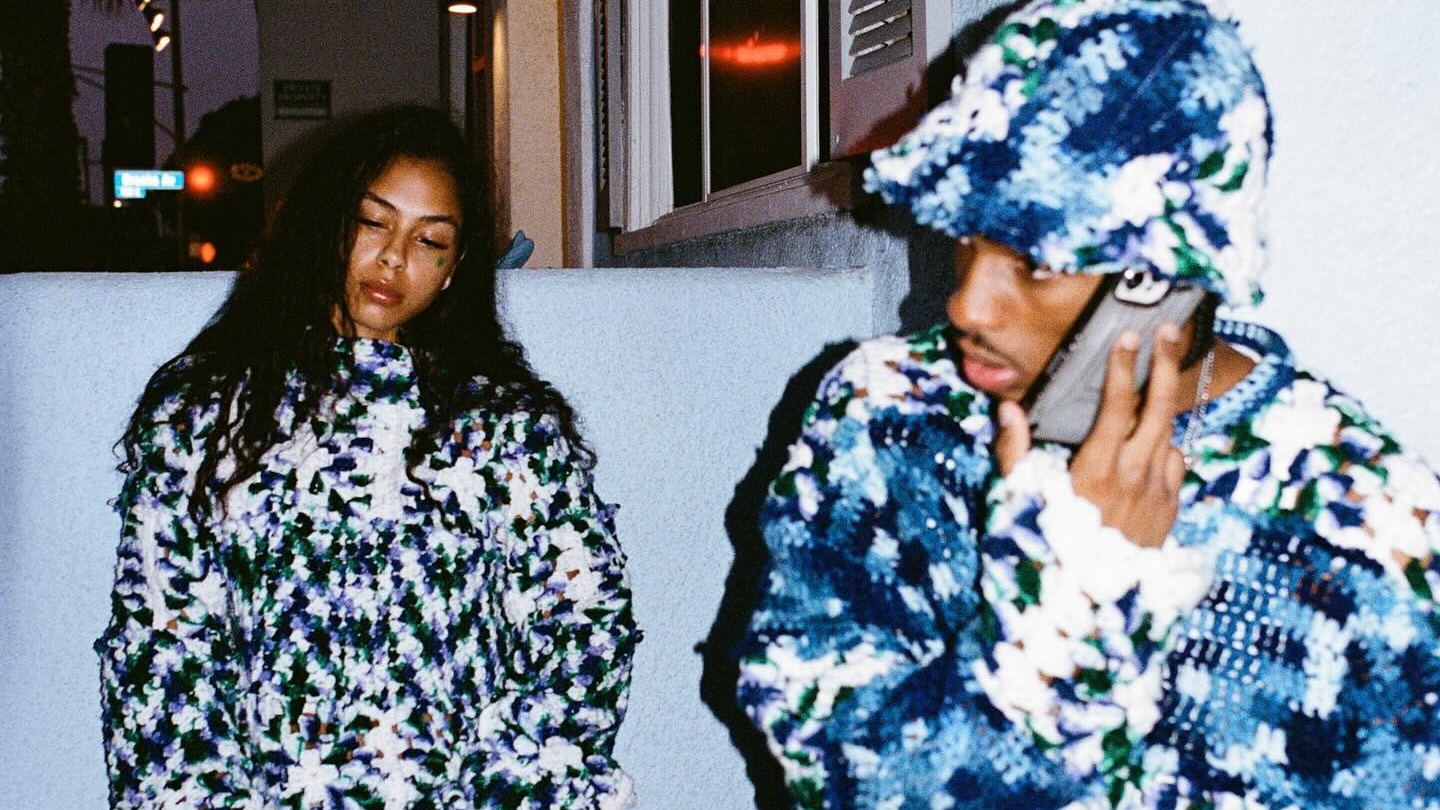 Two people wearing matching blue and white patterned outfits; one is talking on a phone, the other is leaning against a wall. Nigel Xavier