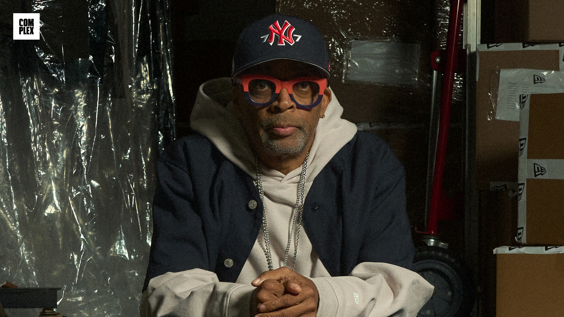 Spike Lee wearing a cap and glasses sits on the back of a truck surrounded by boxes and equipment, in a casual setting.