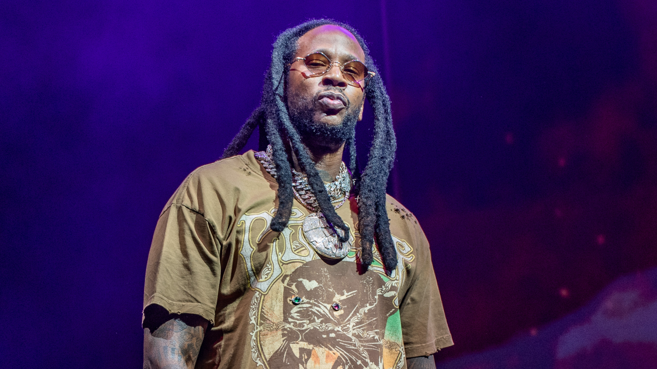 Rapper 2 Chainz on stage, wearing sunglasses and a graphic t-shirt, with a purple-lit background.