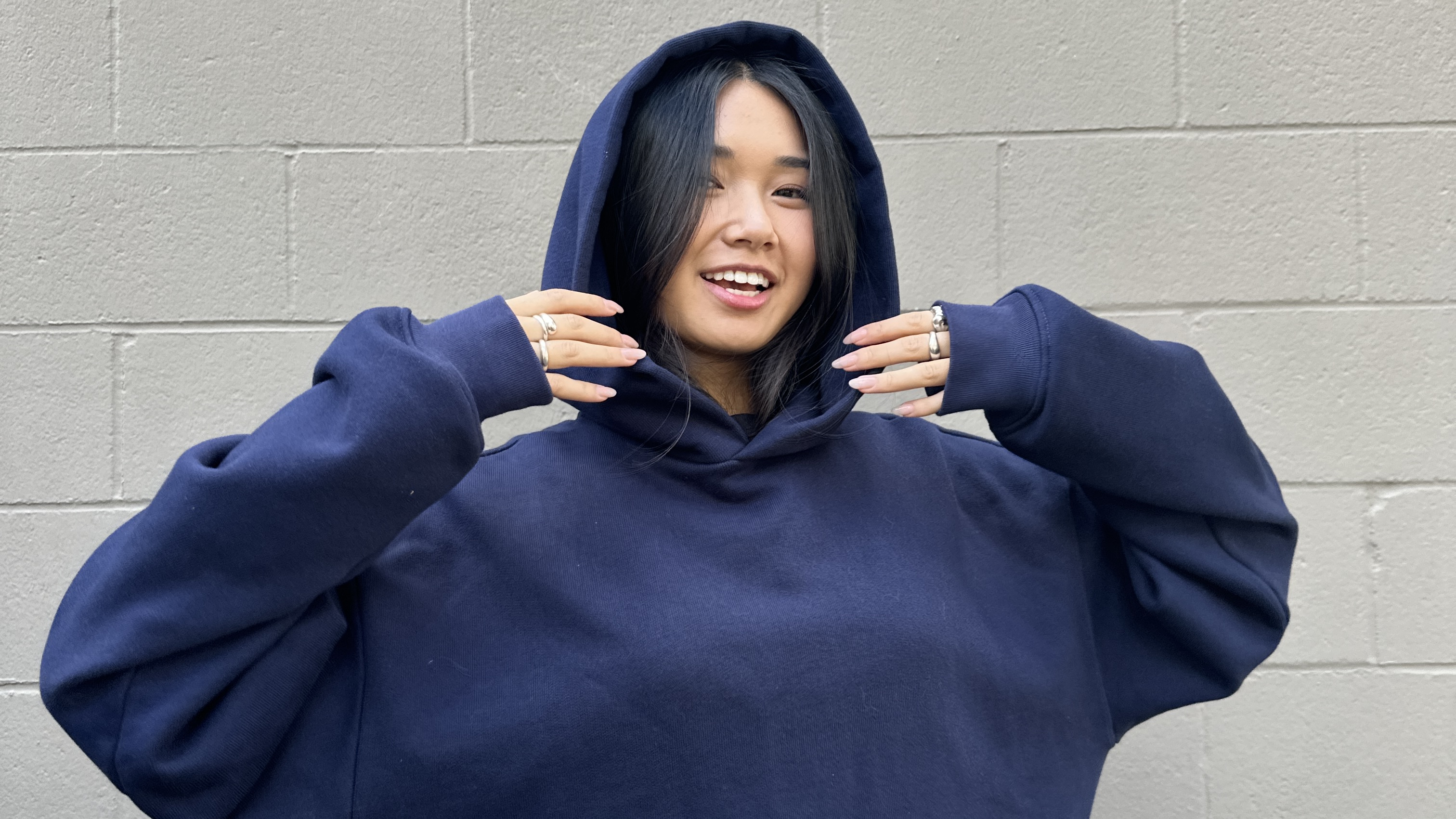 A person in a navy hoodie smiles, standing against a light gray brick wall. They have long dark hair and are wearing rings.