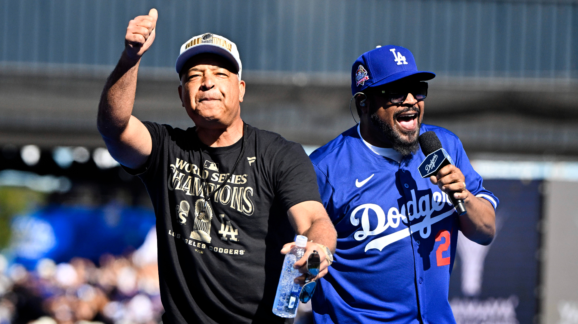 Dave Roberts and Ice Cube celebrating at a Dodgers event. Roberts wears a "World Series Champions" shirt, Ice Cube has a Dodgers jersey and cap on, holding a mic.