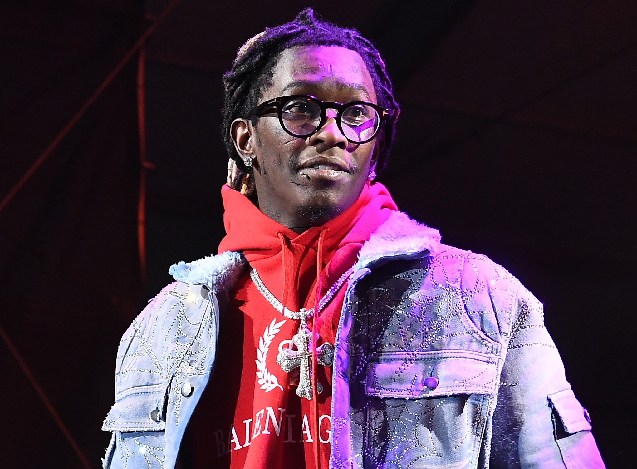 Young Thug on stage wearing glasses, a red Balenciaga hoodie, and a denim jacket, under purple lighting.