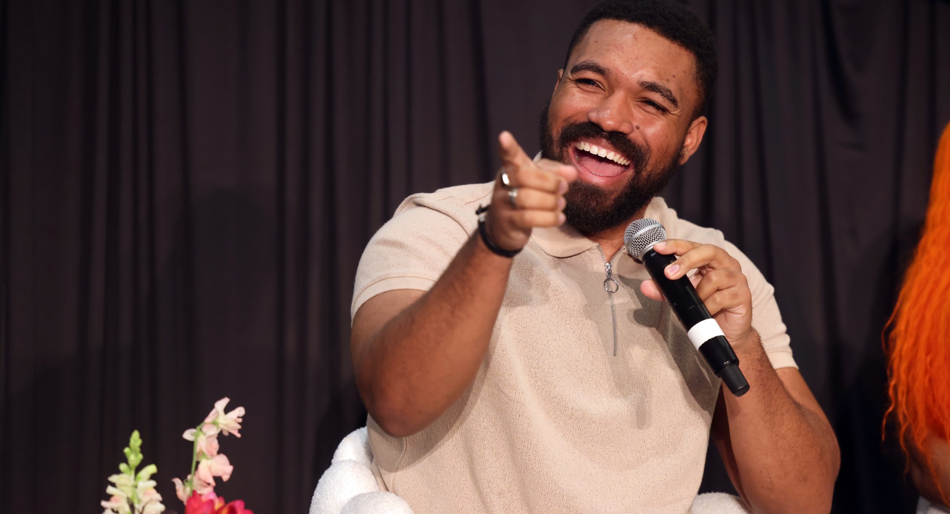 Jordan Howlett holds a microphone while laughing and pointing at something off-screen with flowers in the foreground.