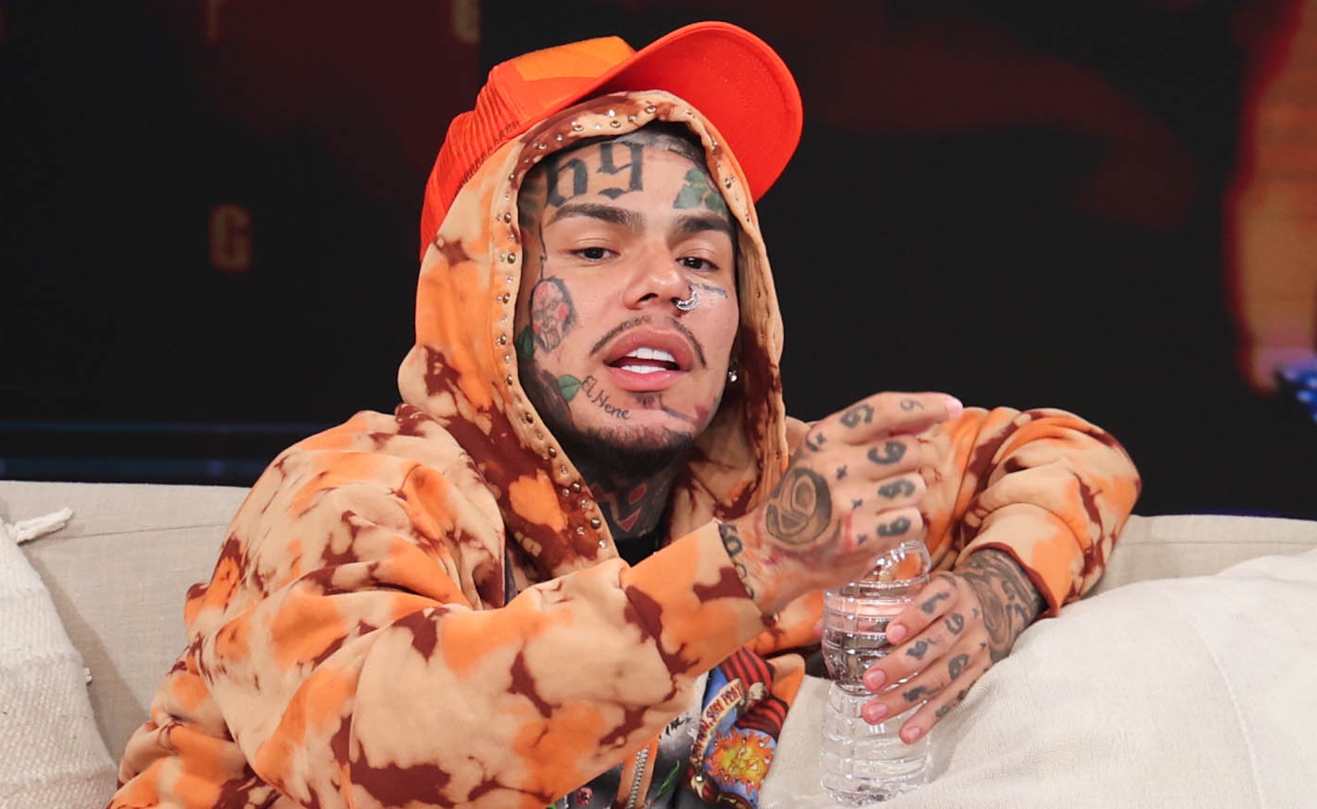 6ix9ine wearing orange hoodie and orange cap while holding plastic water bottle.