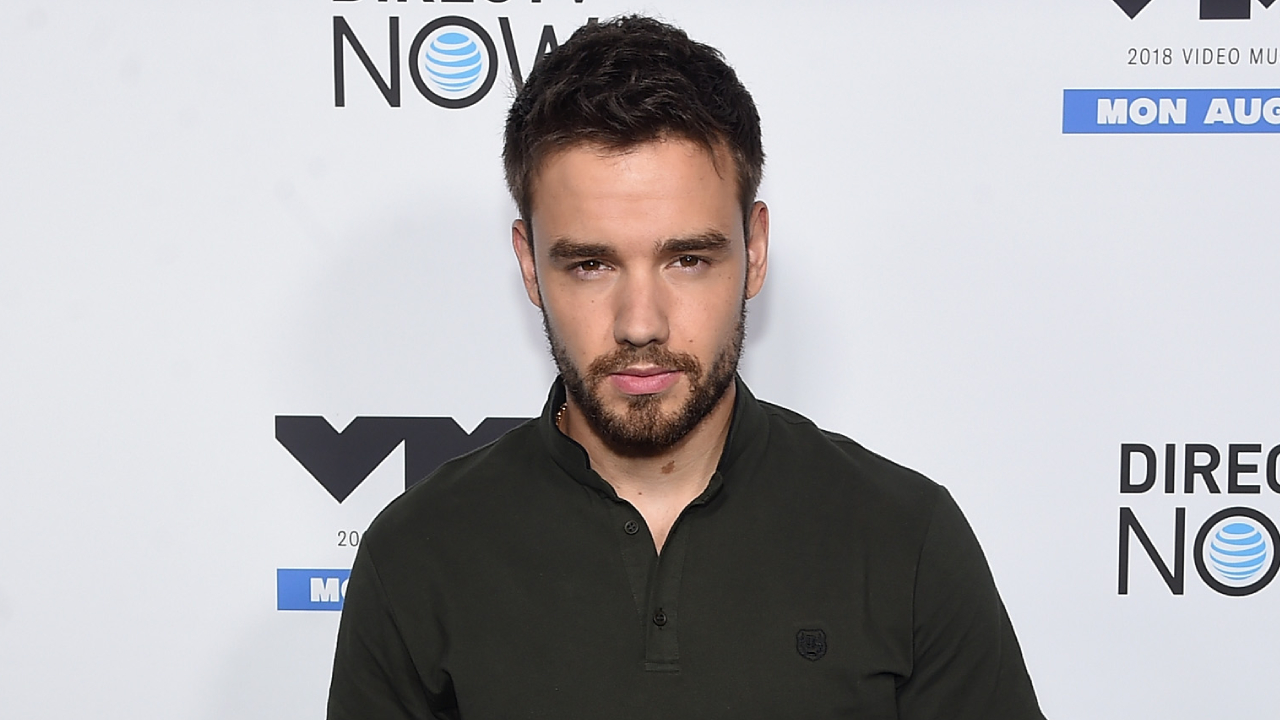 Liam Payne at an event, wearing a dark shirt, standing in front of a promotional backdrop.