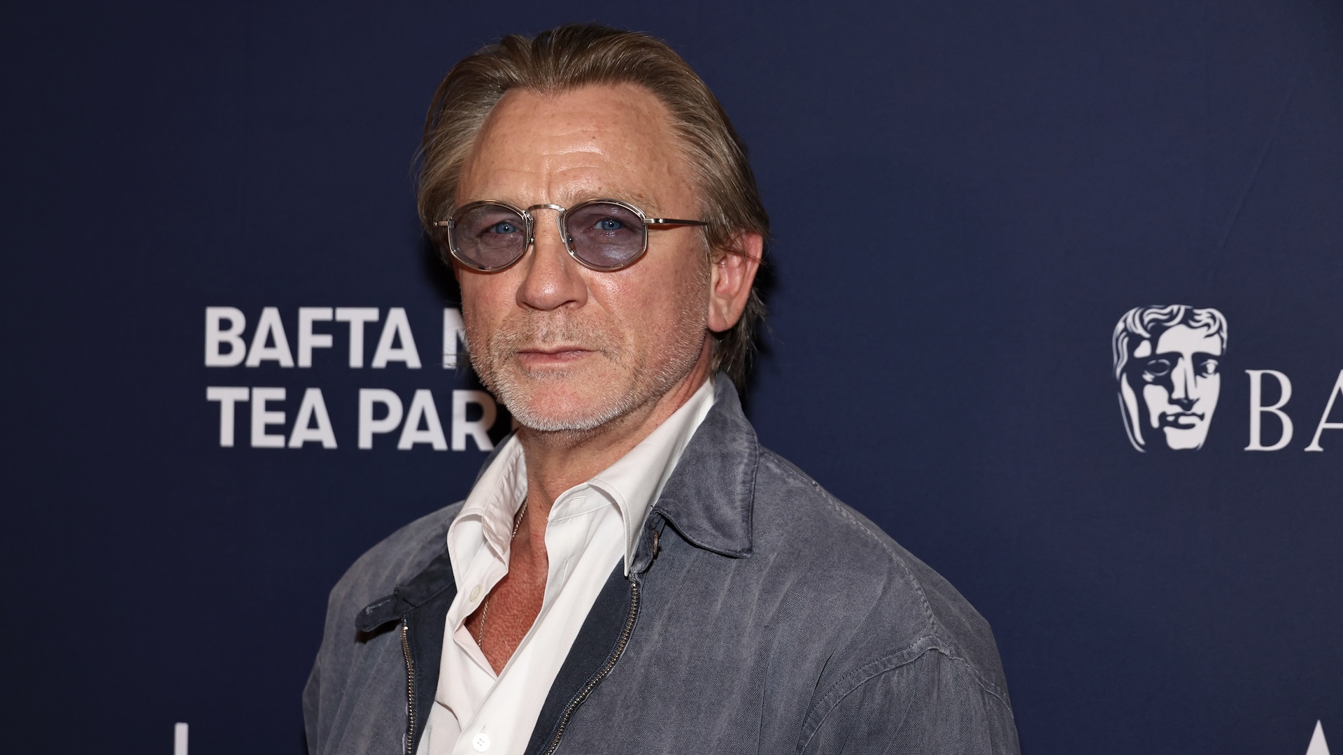 Daniel Craig at a BAFTA event, wearing sunglasses and a casual jacket over a white shirt, posing against a dark backdrop.