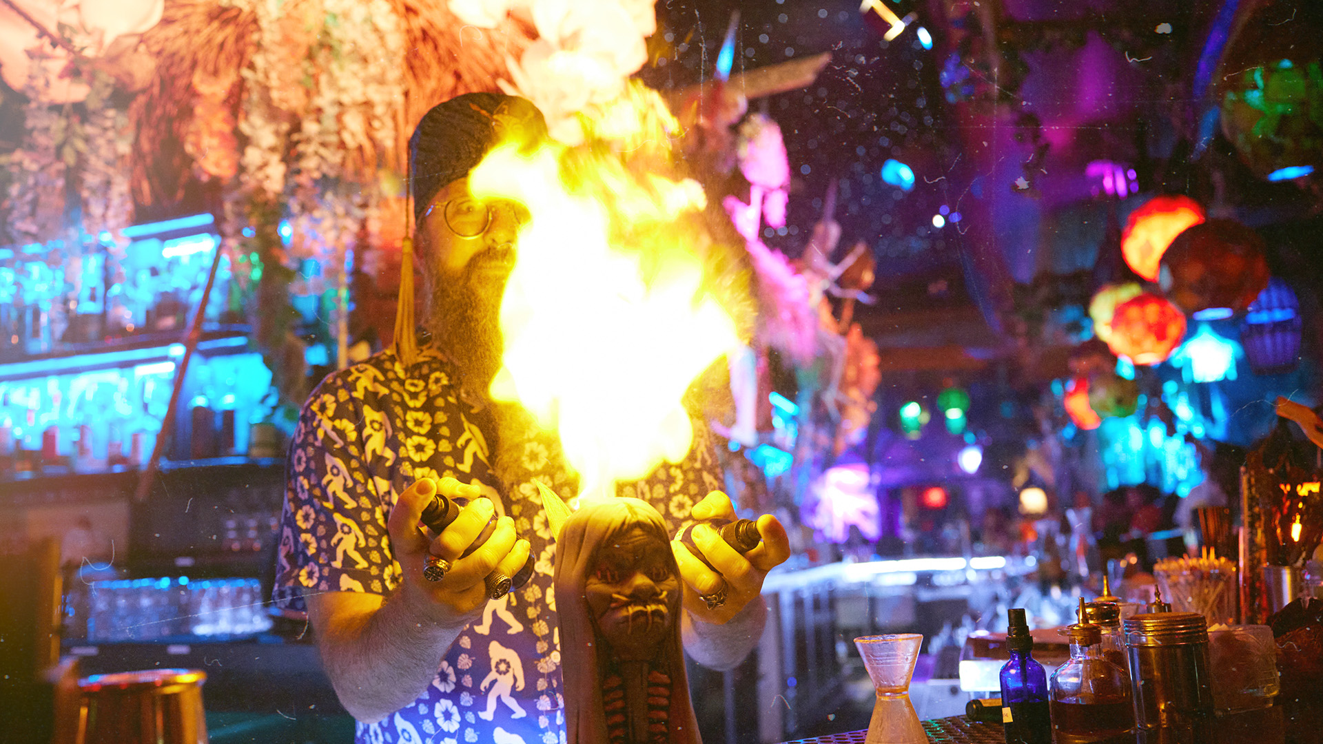 A bartender with a beard and glasses performs a fire trick at a vibrant, colorful bar with tropical decor and various bottles on the counter.