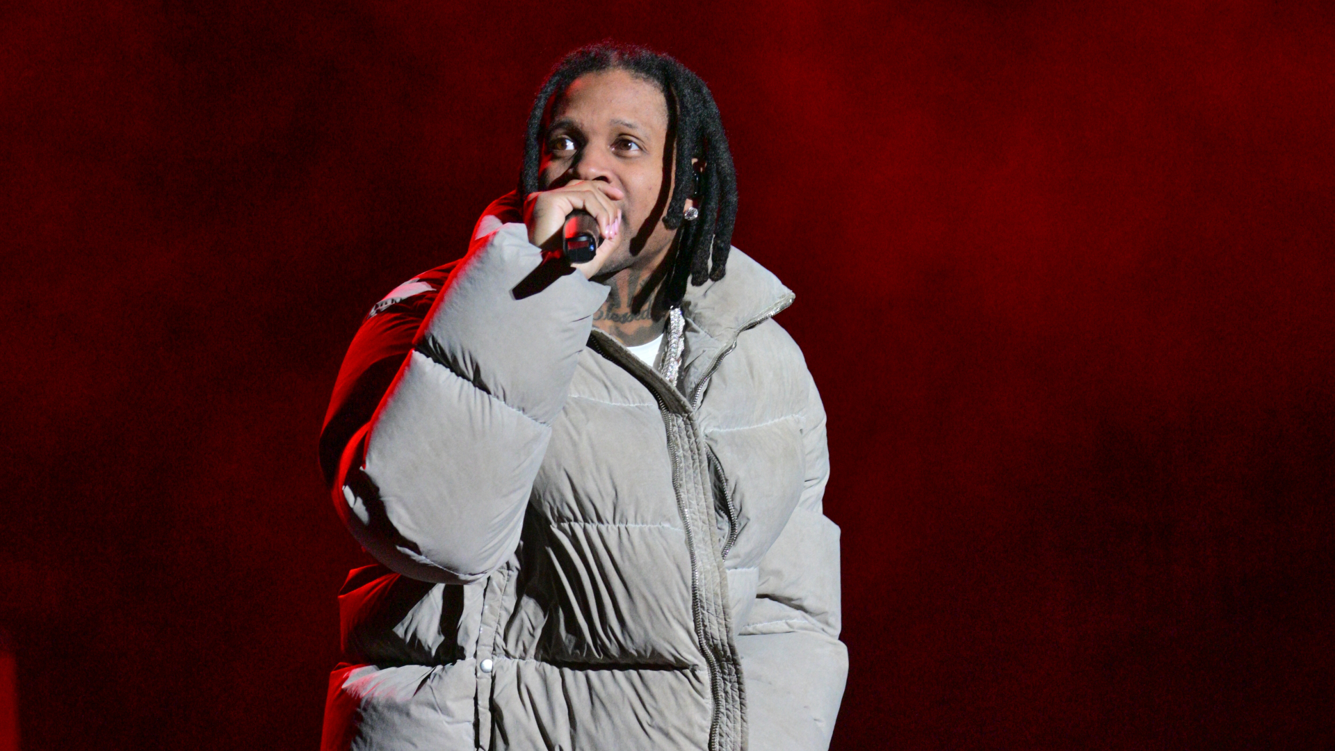 A person wearing a large puffer jacket is performing on stage, holding a microphone, with a red background.
