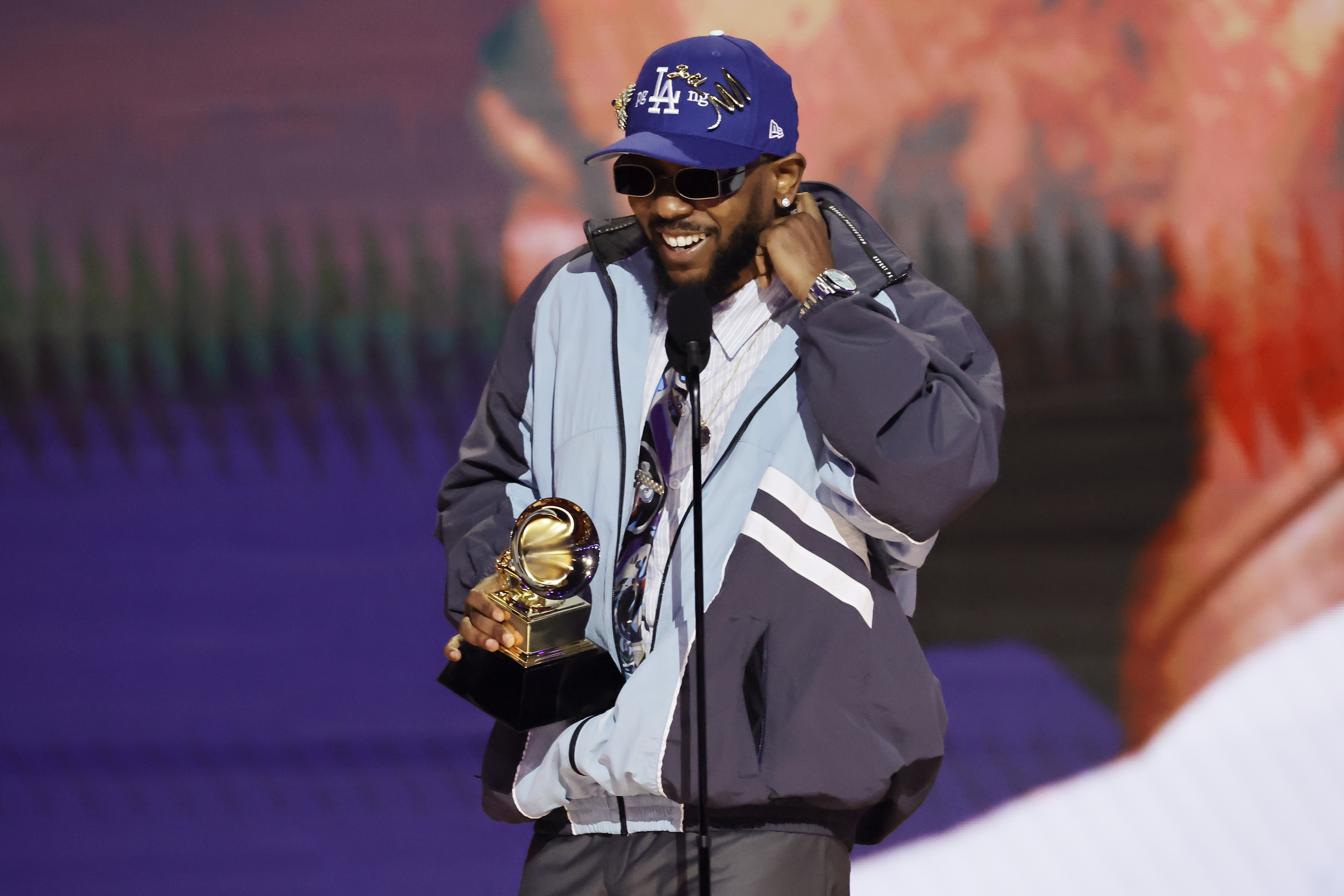 Kendrick Lamar wearing a blue LA Dodgers cap and sunglasses holds a Grammy award on stage, smiling.