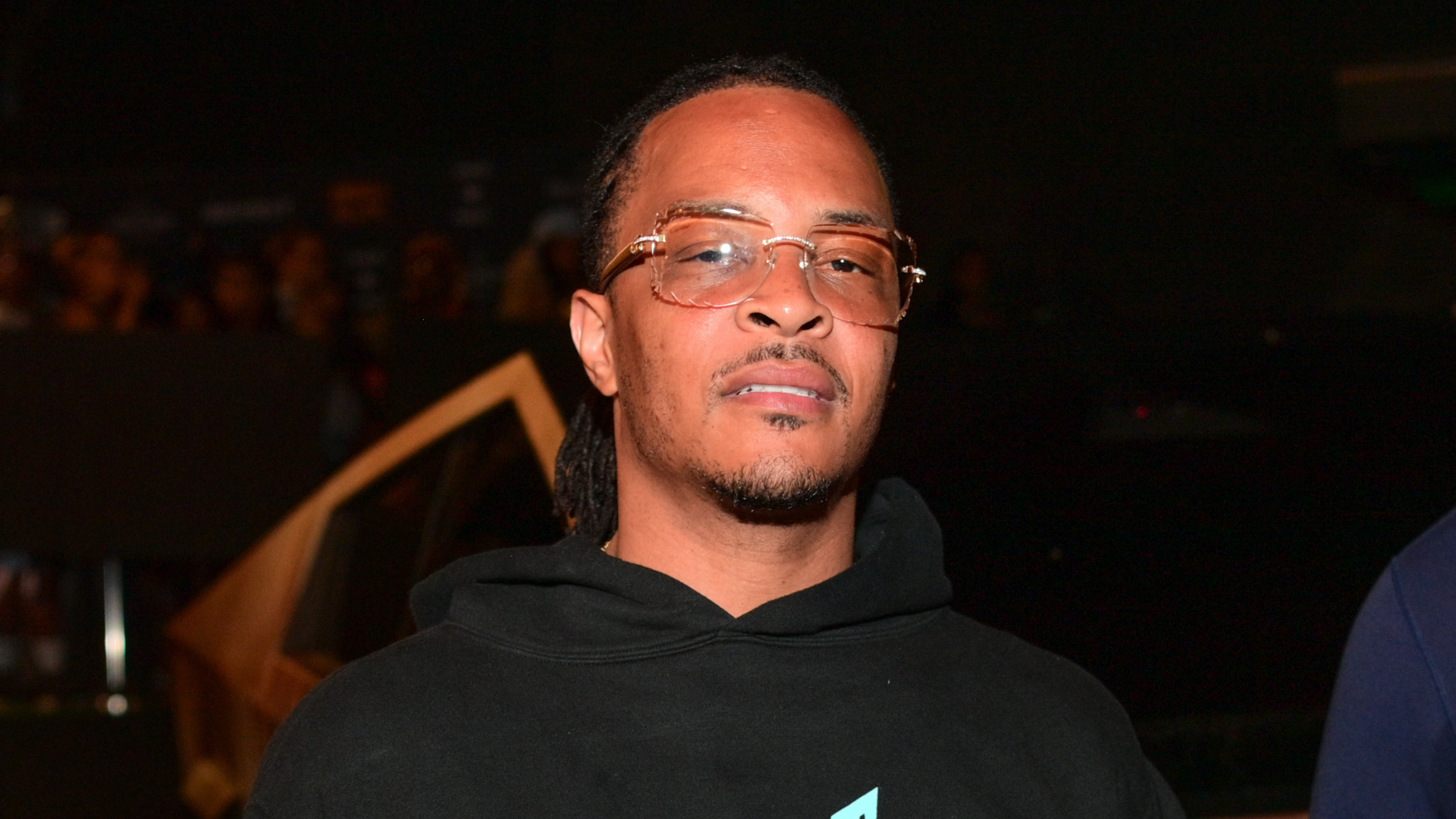 A man wearing glasses and a black hoodie stands in a dimly lit environment, looking towards the camera.