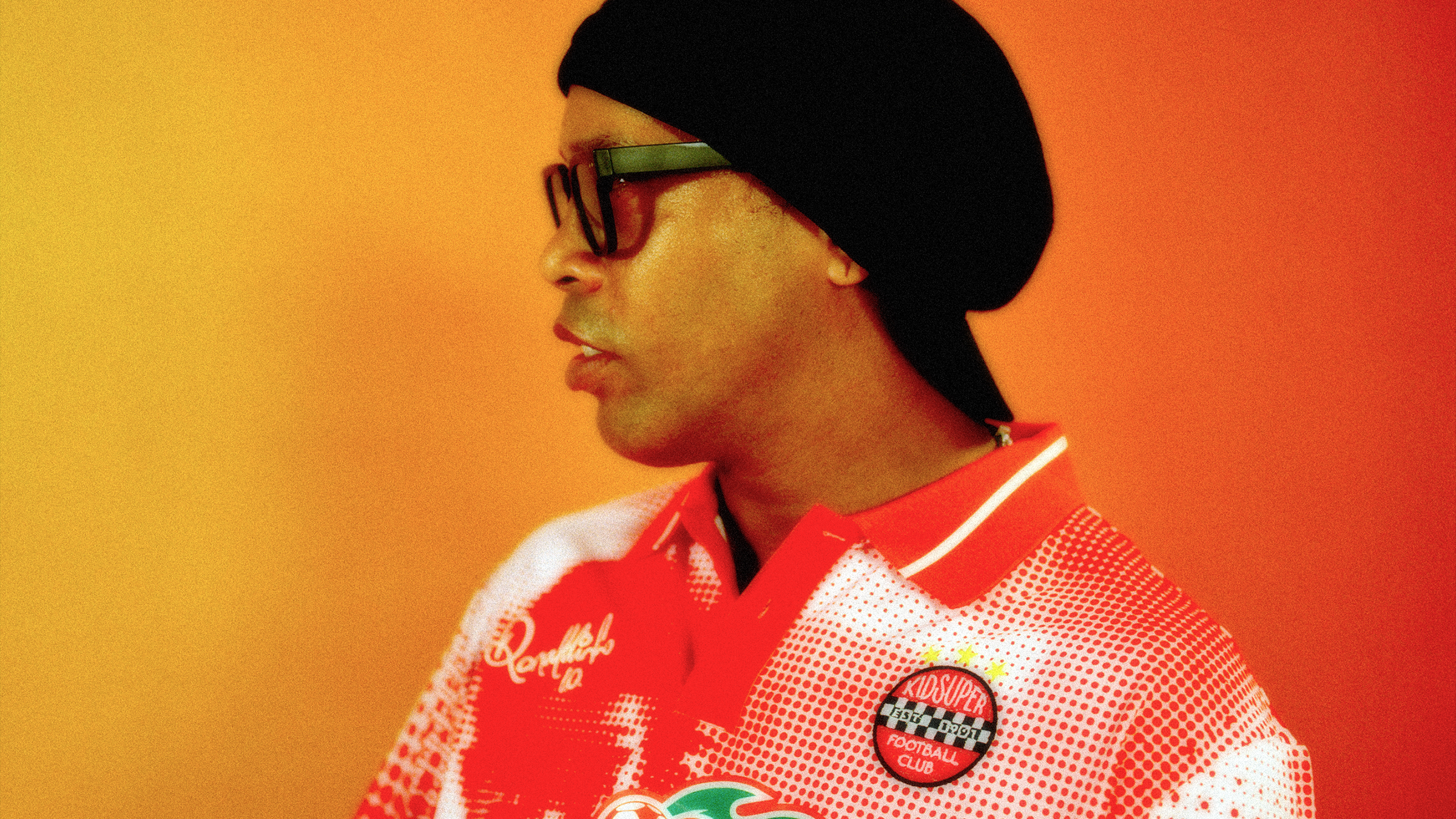 A person in a red and white sports jersey with glasses and a black headscarf, set against a warm orange background.