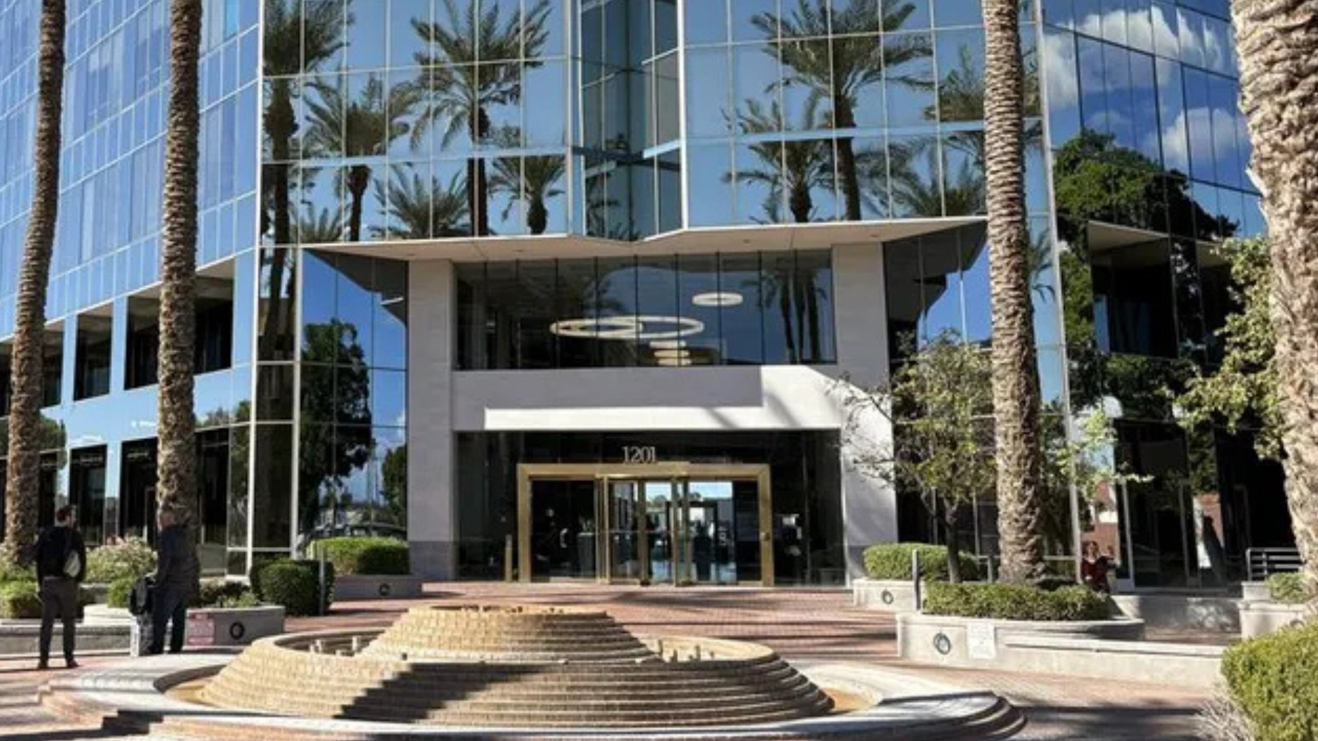 Modern glass building with palm trees and a tiered fountain in front. Reflections of the sky and trees are visible on the windows.