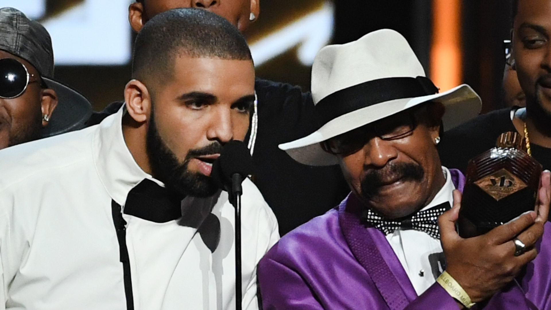 Drake, in a white tuxedo, speaks at a microphone. Beside him, a man in a purple suit and white hat holds an award.