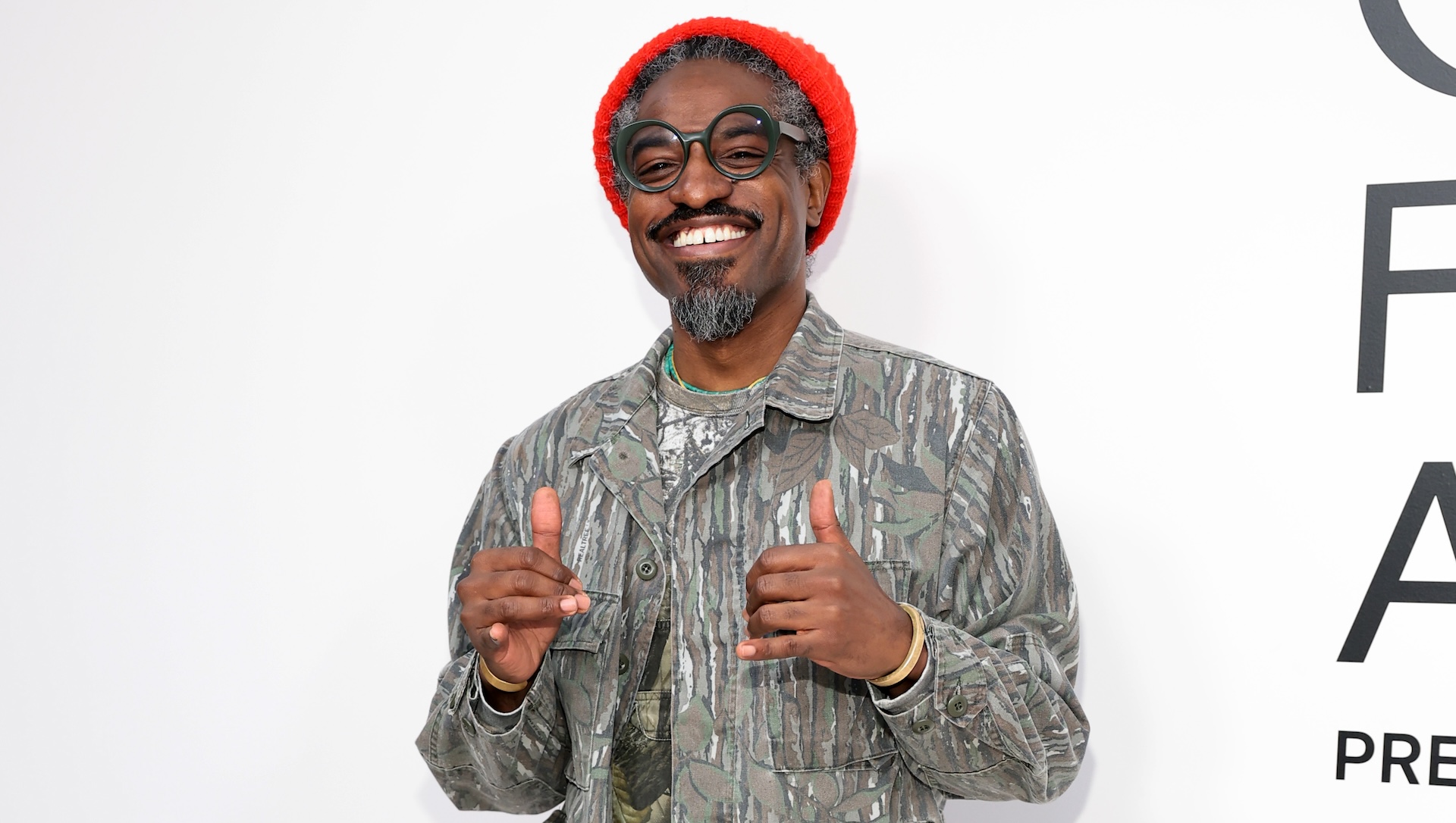 Andre 3000 wearing a red beanie and patterned jacket, smiling and giving thumbs up.