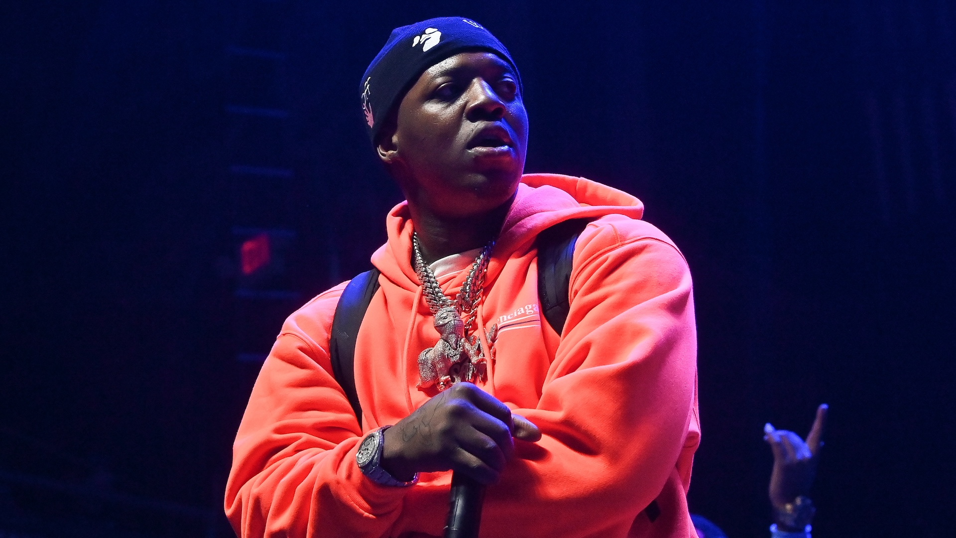 Lil Zay Osama performs on stage, holding a microphone. He wears several chains and a watch.