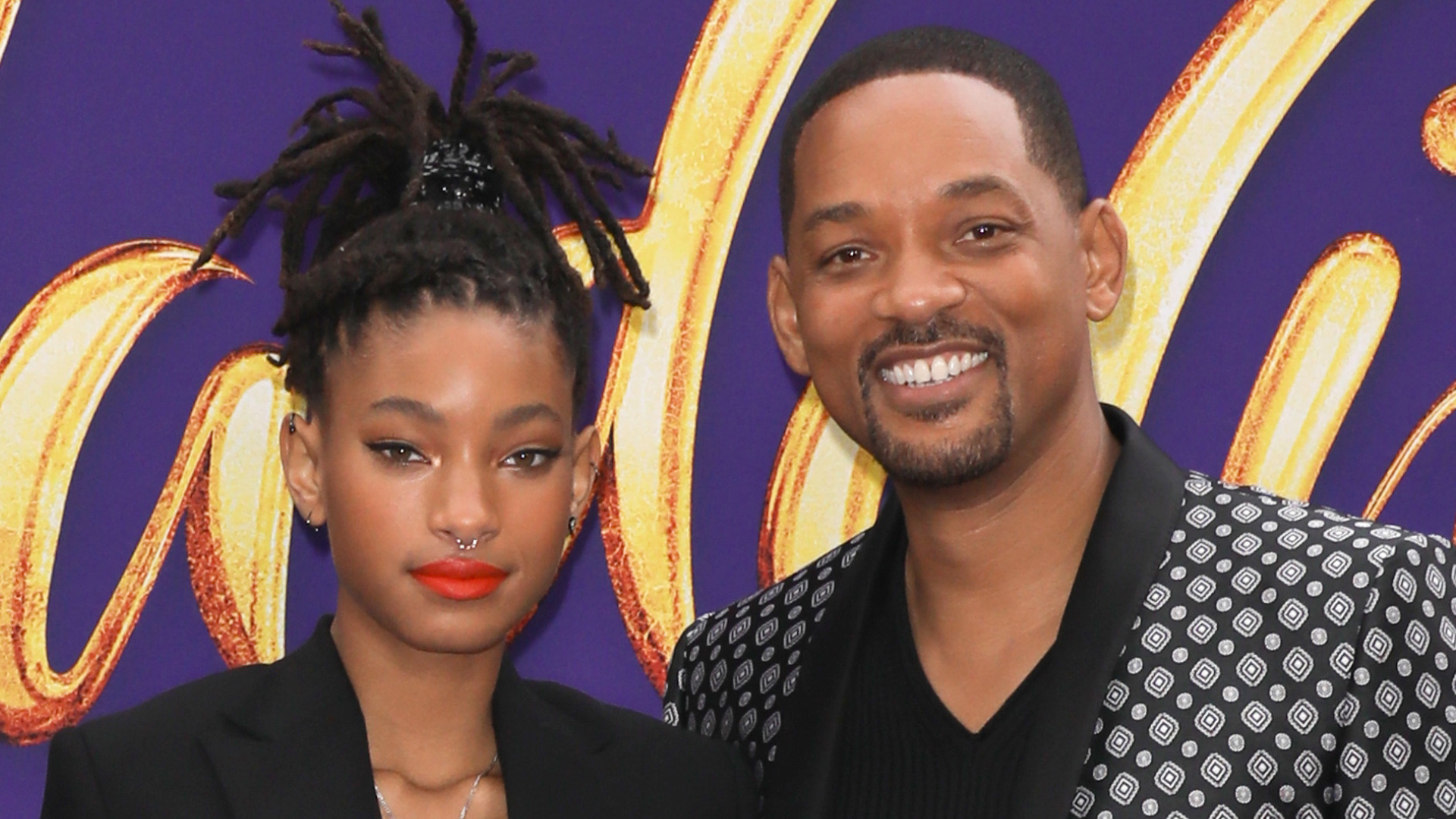 Willow Smith and Will Smith smiling at an event, with a vibrant purple and gold background.