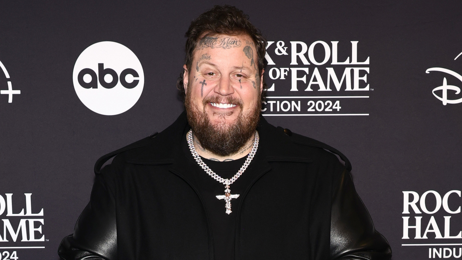 A man with face tattoos and a beard, wearing a black jacket and a chain necklace, stands in front of a Rock & Roll Hall of Fame backdrop.