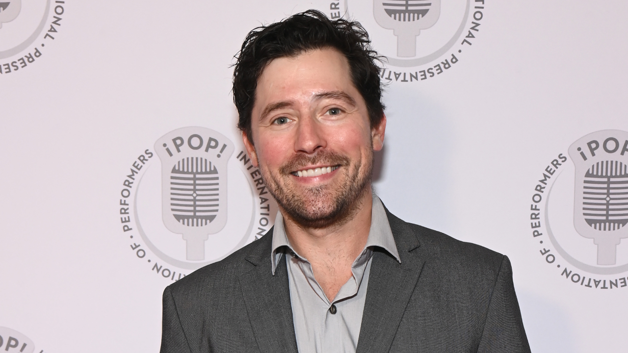 Robert Vito in a gray suit smiles at an event with a backdrop featuring a microphone logo and the text "iPOP! International Presentation of Performers."