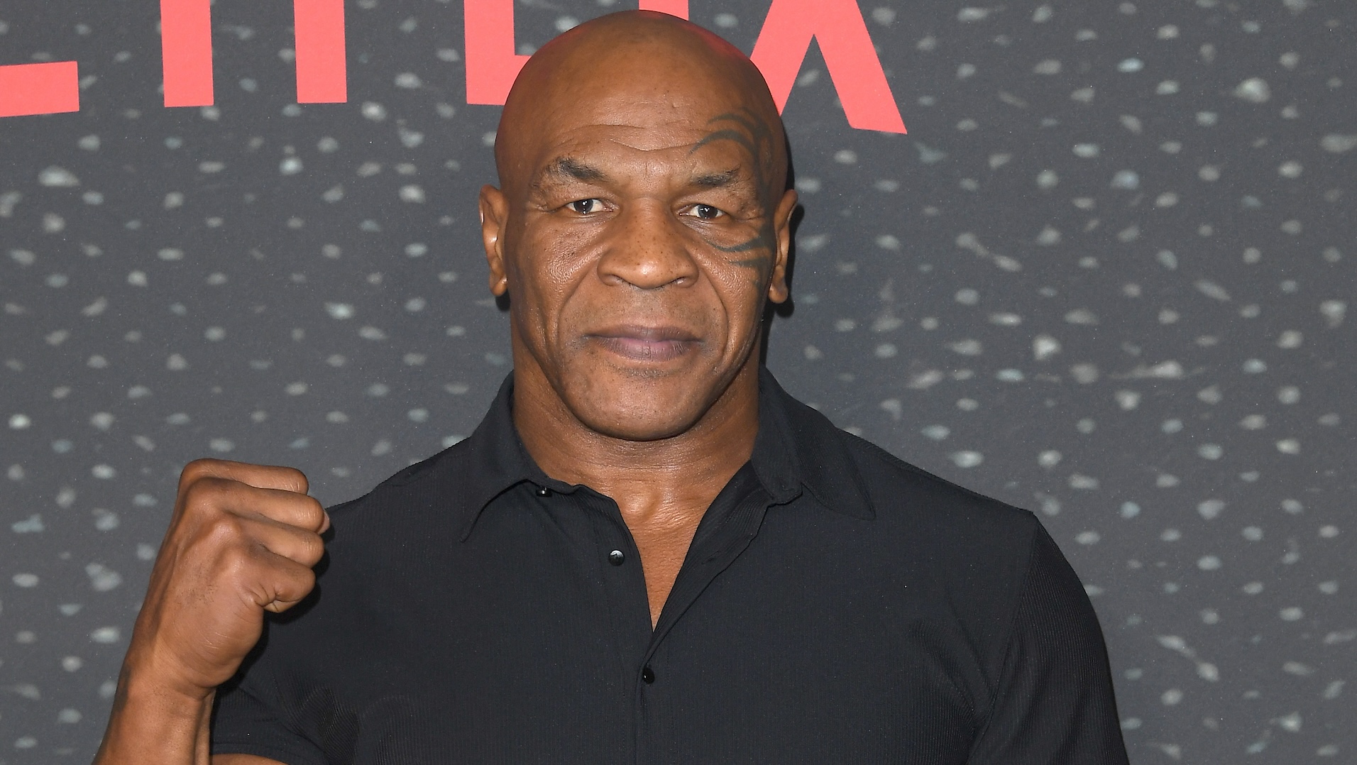 Mike Tyson, wearing a black shirt, poses with a fist raised. Background features a Netflix logo.