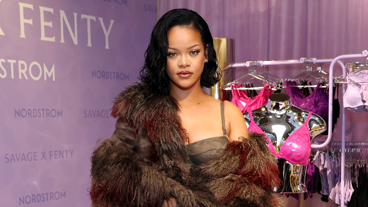 Rihanna at a Savage X Fenty event, wearing a fur coat. Mannequins with lingerie are displayed in the background.
