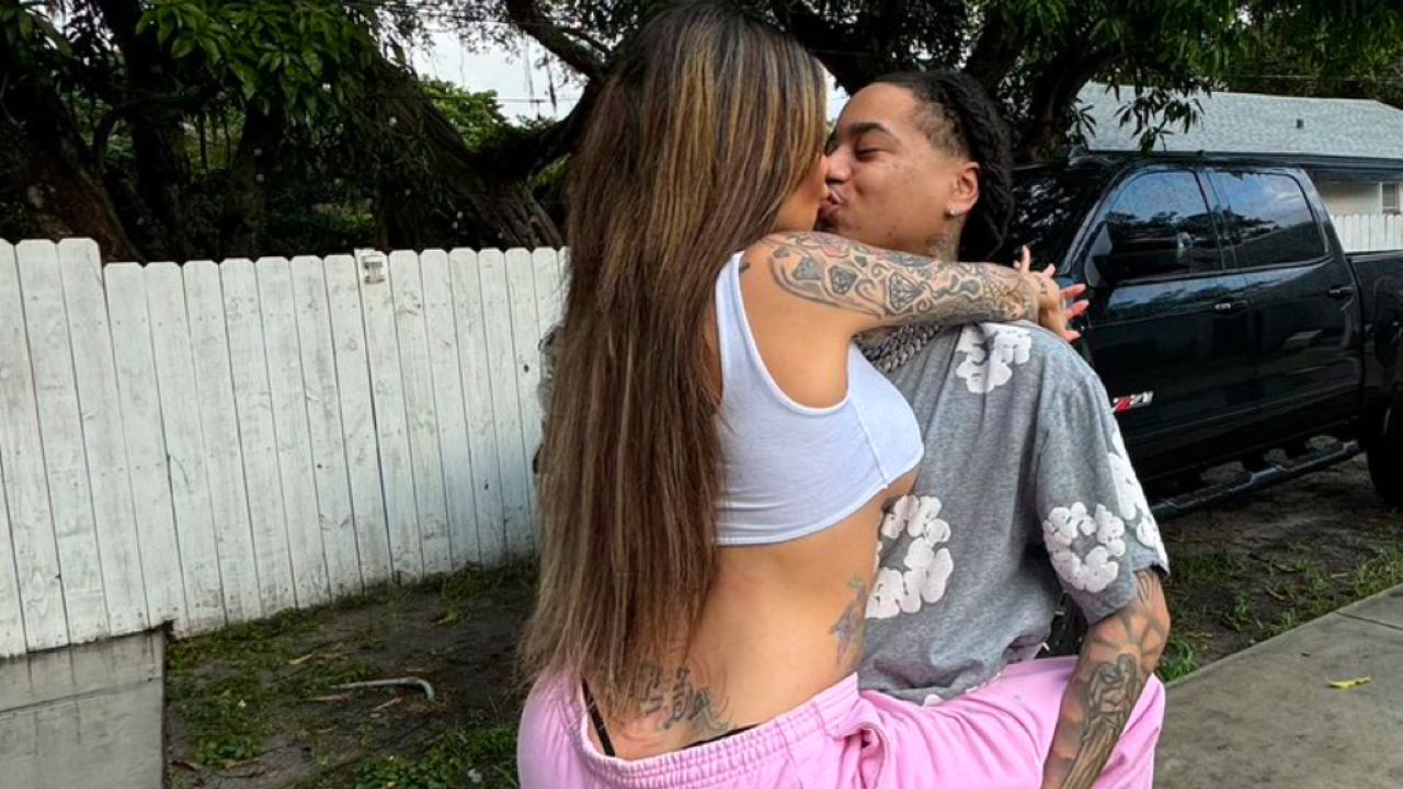 A couple embracing and kissing outdoors, with tattoos visible, near a white fence and a black truck.