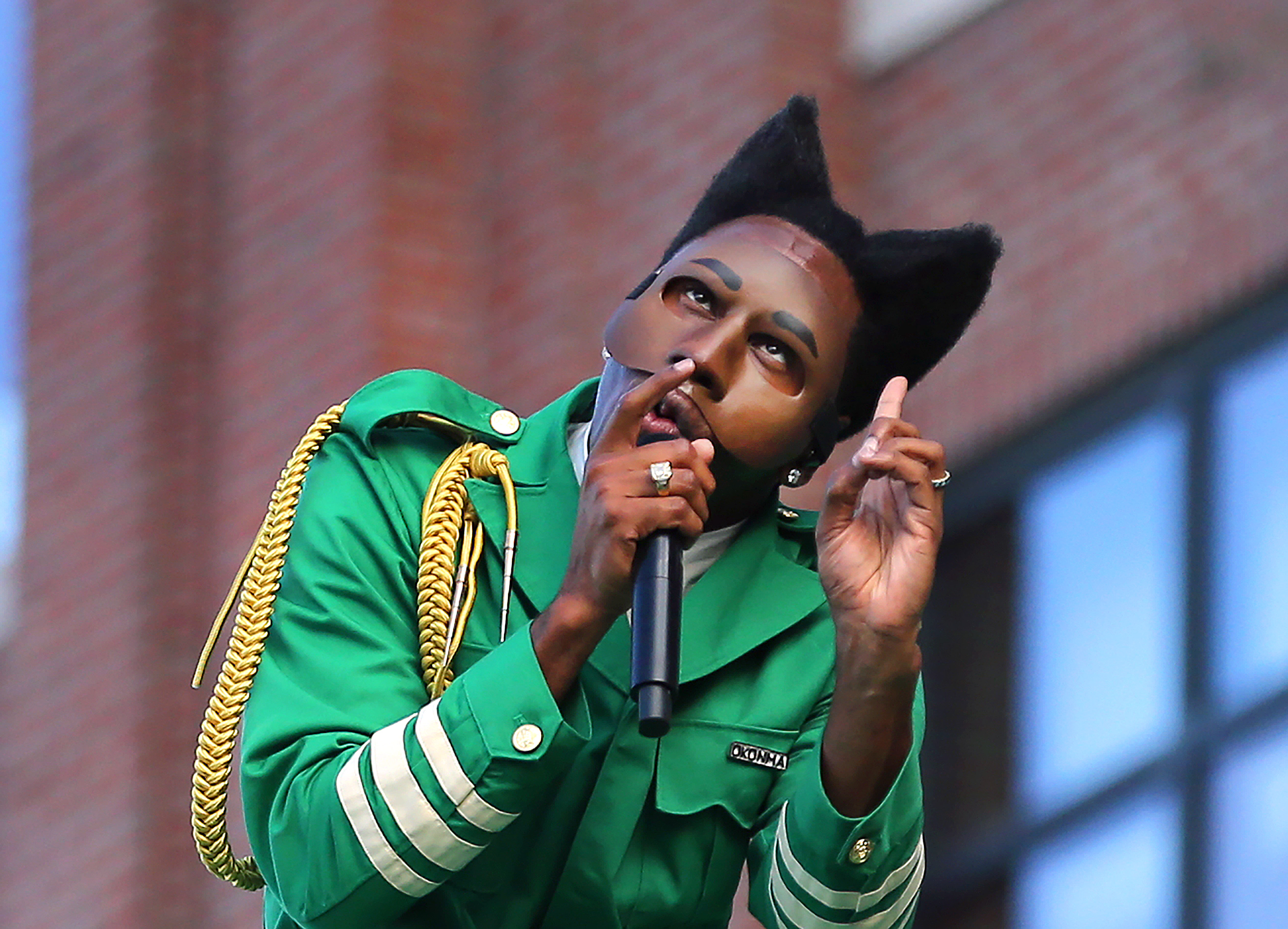 Tyler, the Creator in a green costume with a cat-like mask, holding a microphone, gestures dramatically on stage.