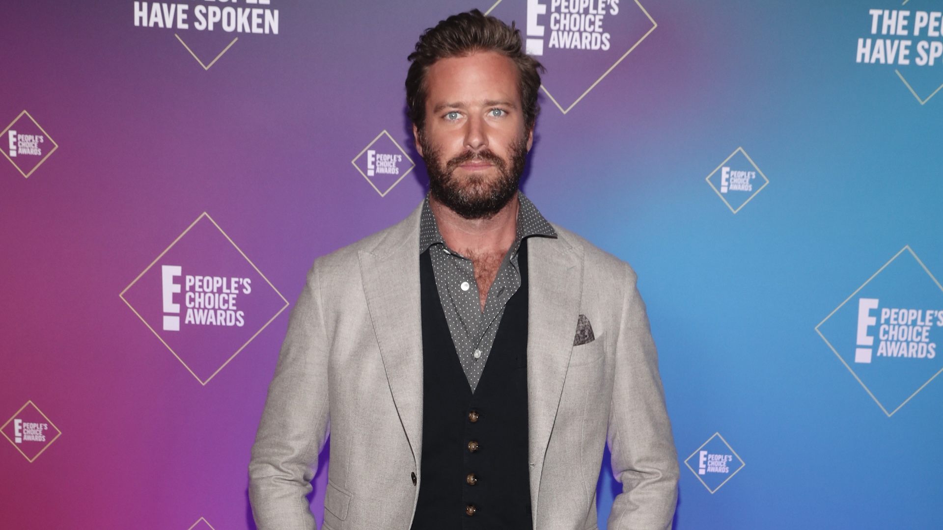 Armie Hammer at the People's Choice Awards, wearing a light gray blazer over a dark vest and shirt, against a colorful backdrop.