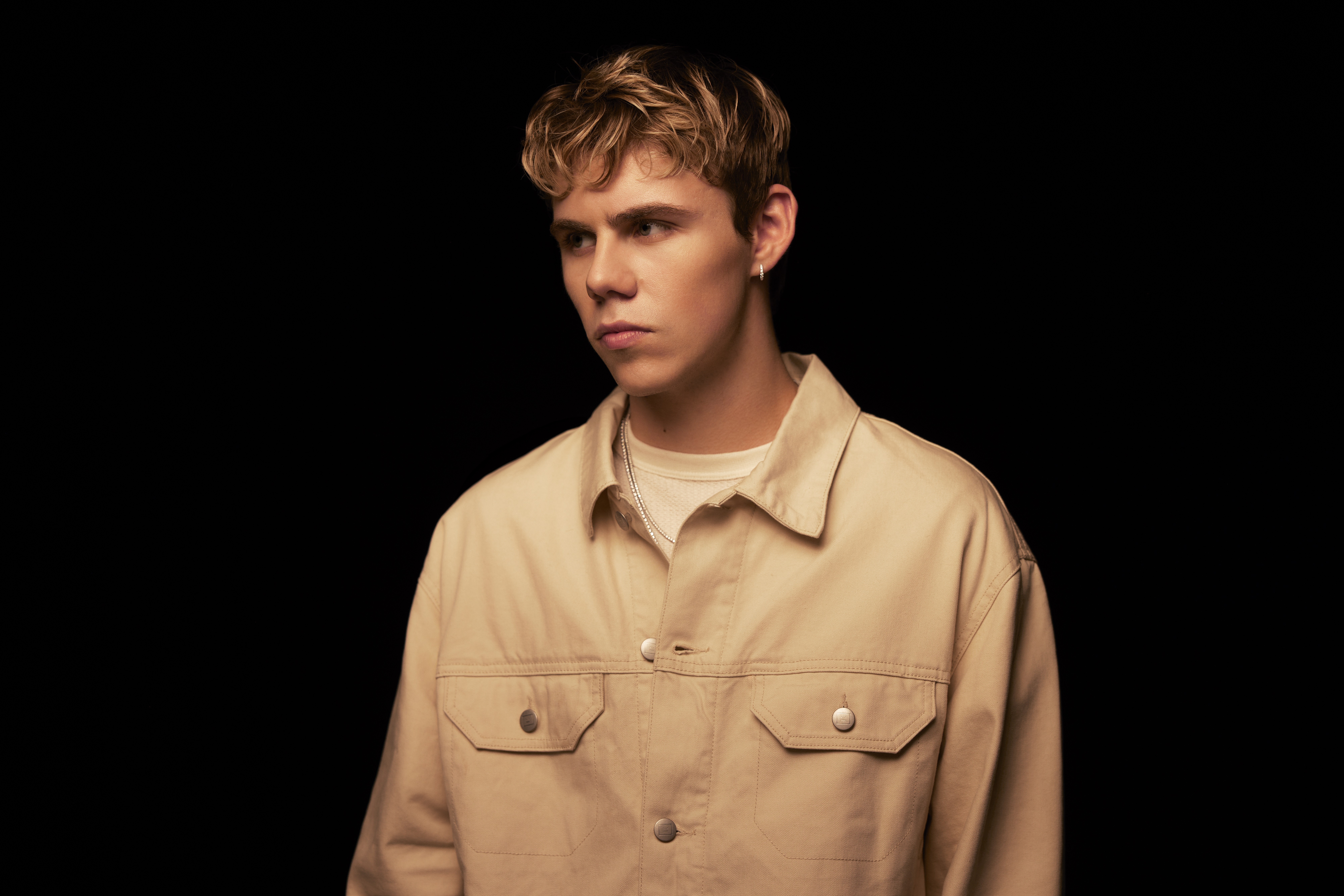 Young man with short, wavy hair wearing a beige jacket, standing against a dark background, looking to the side.