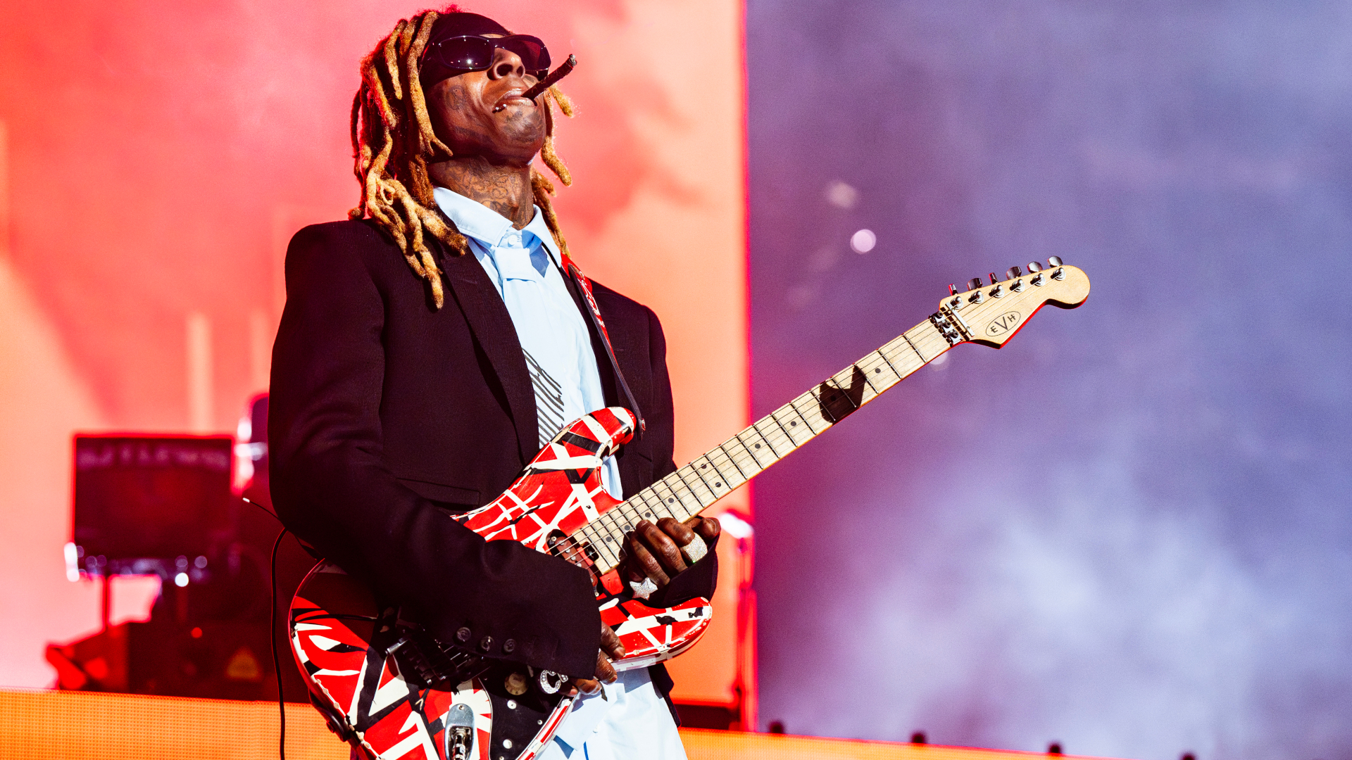 Lil Wayne performing on stage, wearing sunglasses and playing a red and white electric guitar.