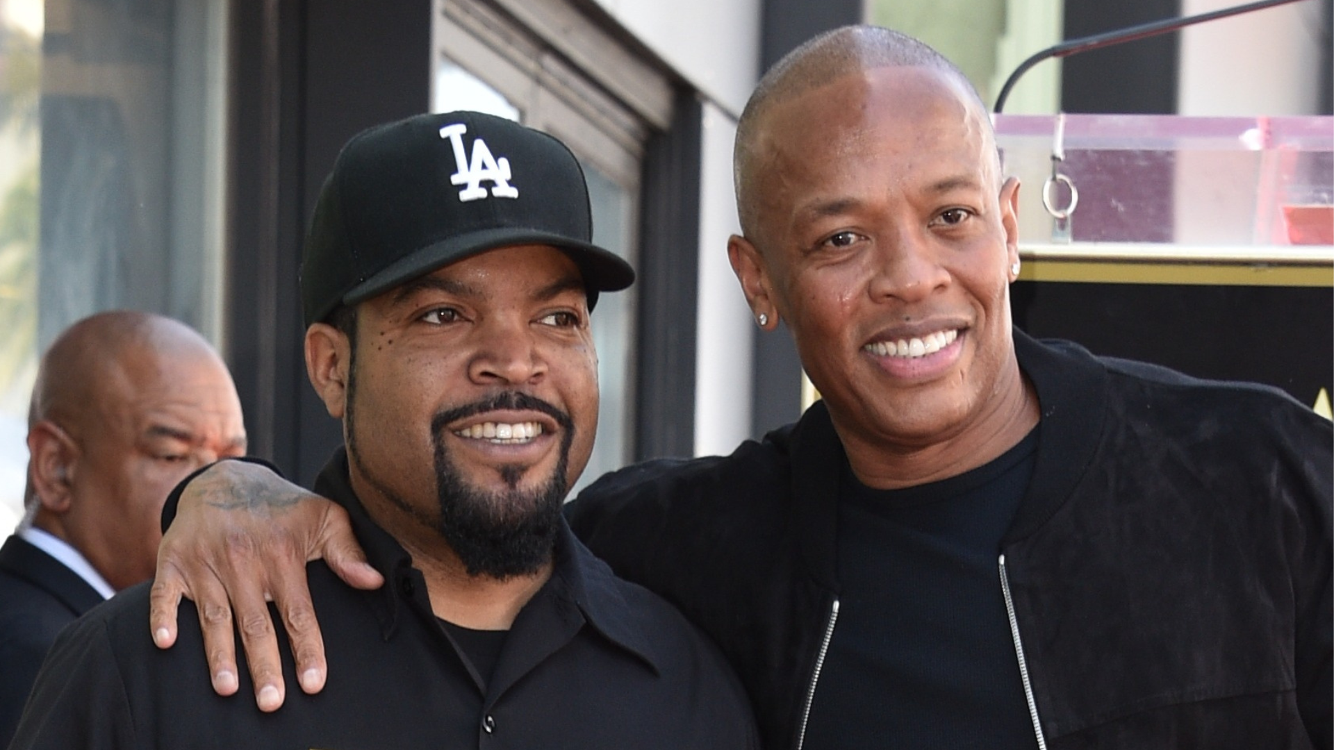 Ice Cube and Dr. Dre smiling together. Ice Cube wears a black LA cap, and Dr. Dre is in a black jacket.