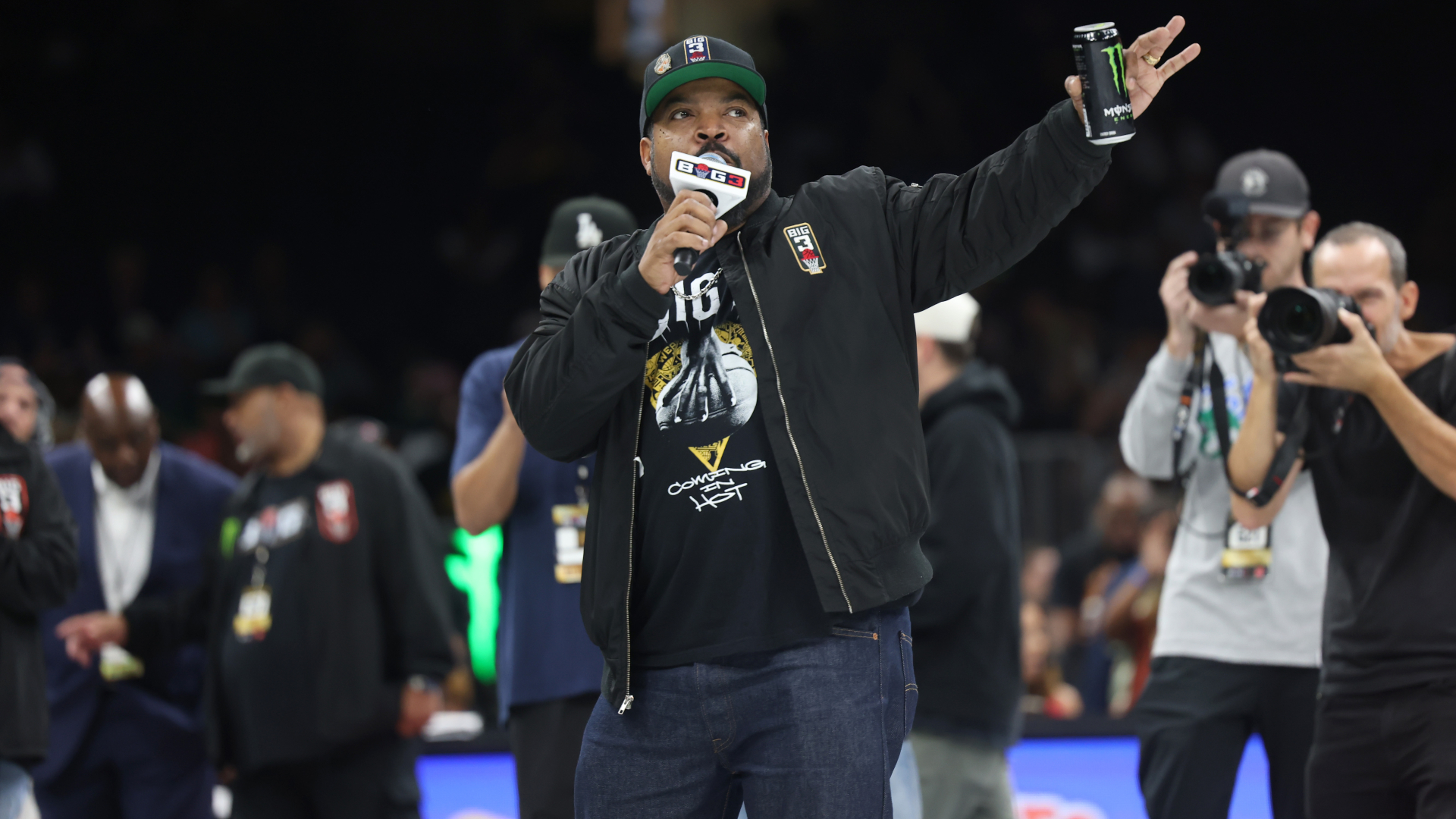 Ice Cube holds a microphone and a can, speaking at an event. He's wearing a cap and jacket, with people and photographers around him.