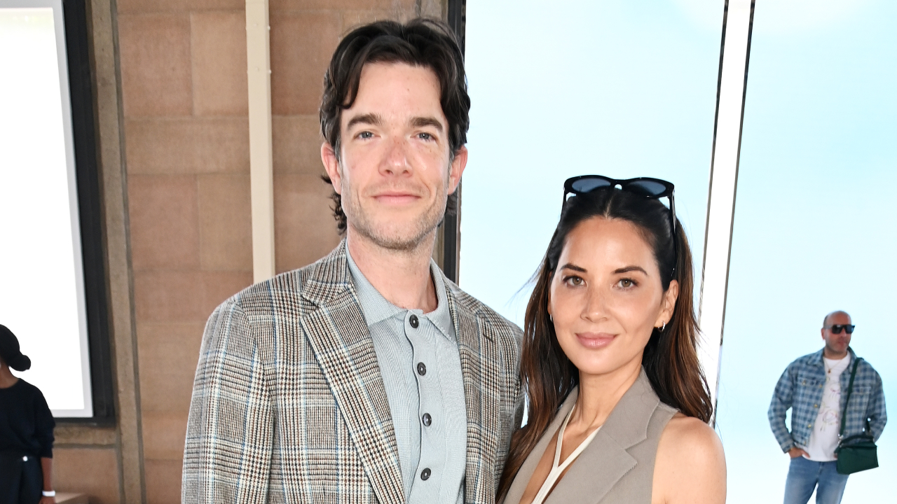 John Mulaney and Olivia Munn posing together, Mulaney in a plaid jacket and Munn in a beige outfit with sunglasses on her head.