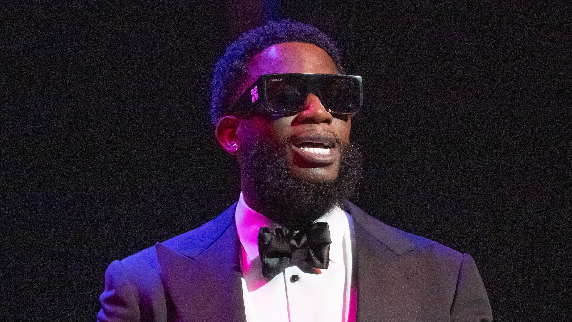 A man in a tuxedo with a bow tie and large sunglasses stands against a dark background.