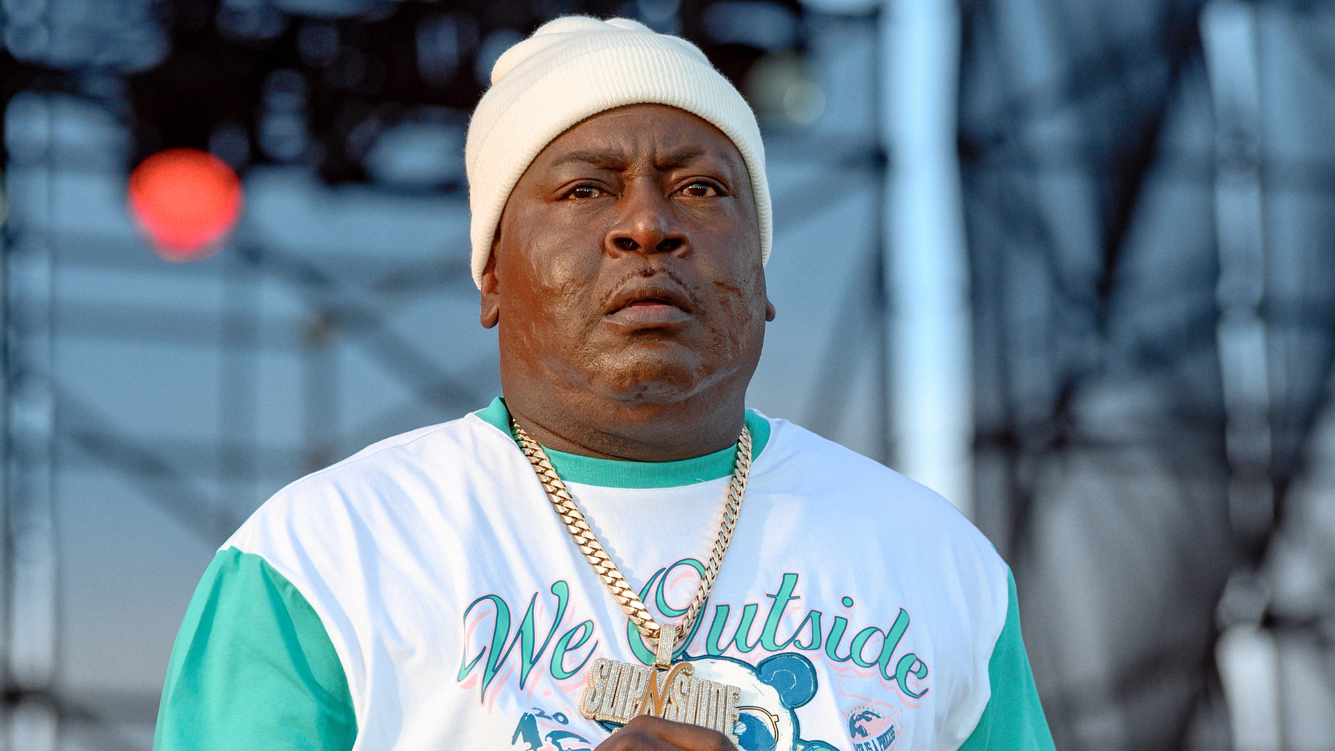 Trick Daddy wears a white beanie, gold chain, and a "We Outside" shirt while standing on stage.