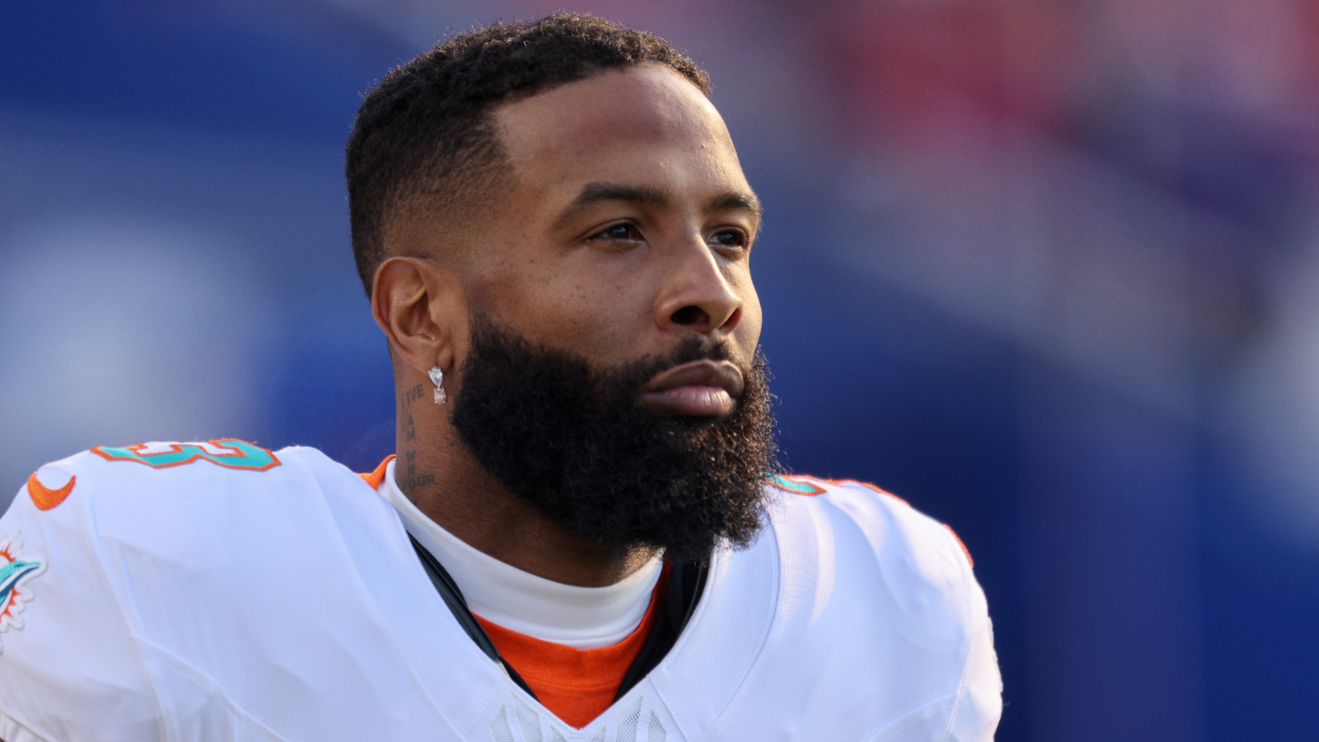 A football player with a beard, wearing a white jersey, looks focused. The background is blurred.