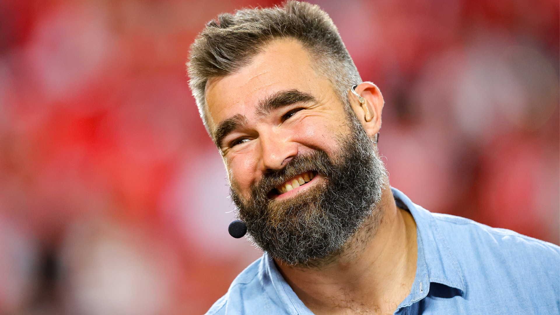 A man with a beard and a headset microphone smiles, wearing a light blue shirt. The background is blurred and red.