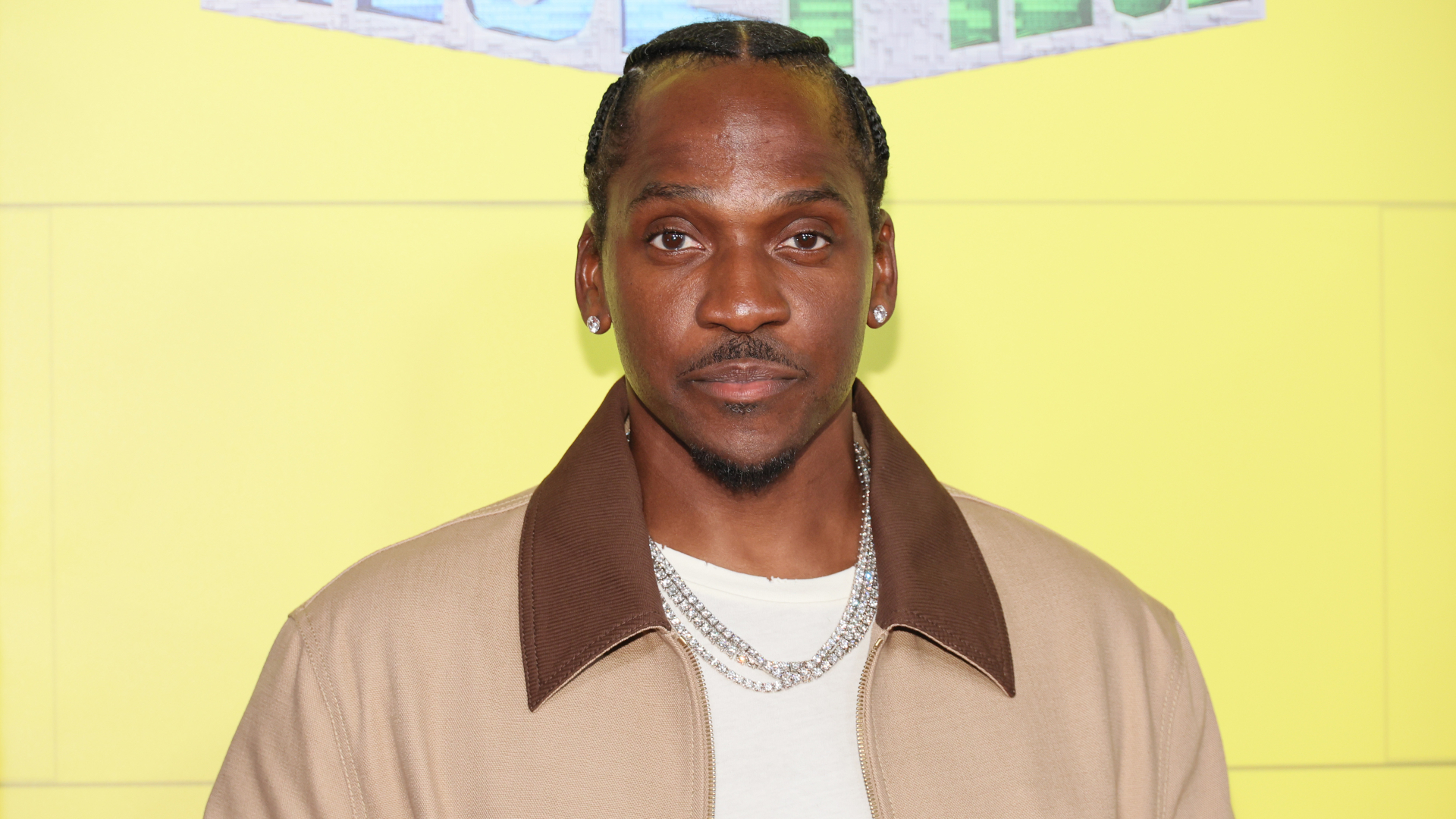 Pusha T at an event, wearing a beige jacket with a white shirt and chain necklace, against a yellow background.